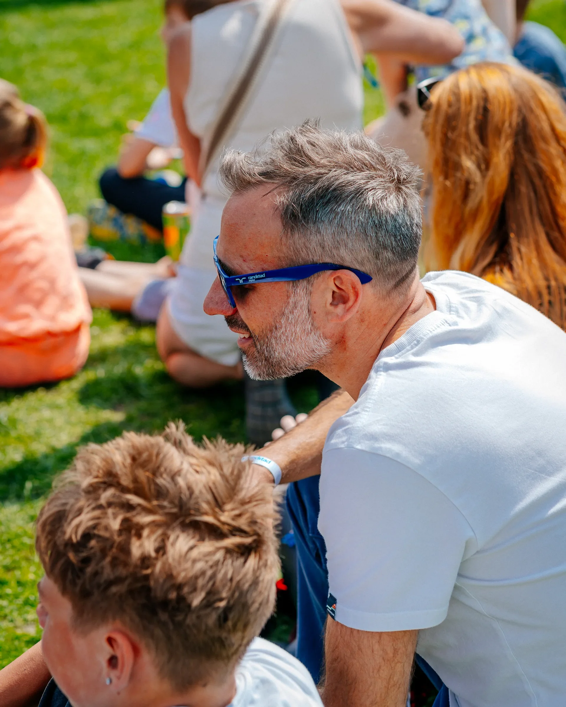 Man met grijze haar en een baard draagt blauwe zonnebril en zit buiten op gras, omringd door mensen en een hond.