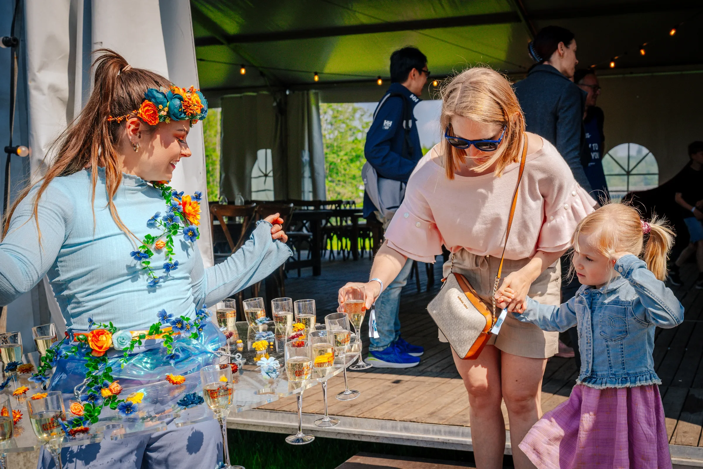 Vrouw met bloemenkroon en bloemenketting deelt drankjes uit aan jonge meisje op een feest, terwijl andere mensen in de achtergrond genieten onder een tent.