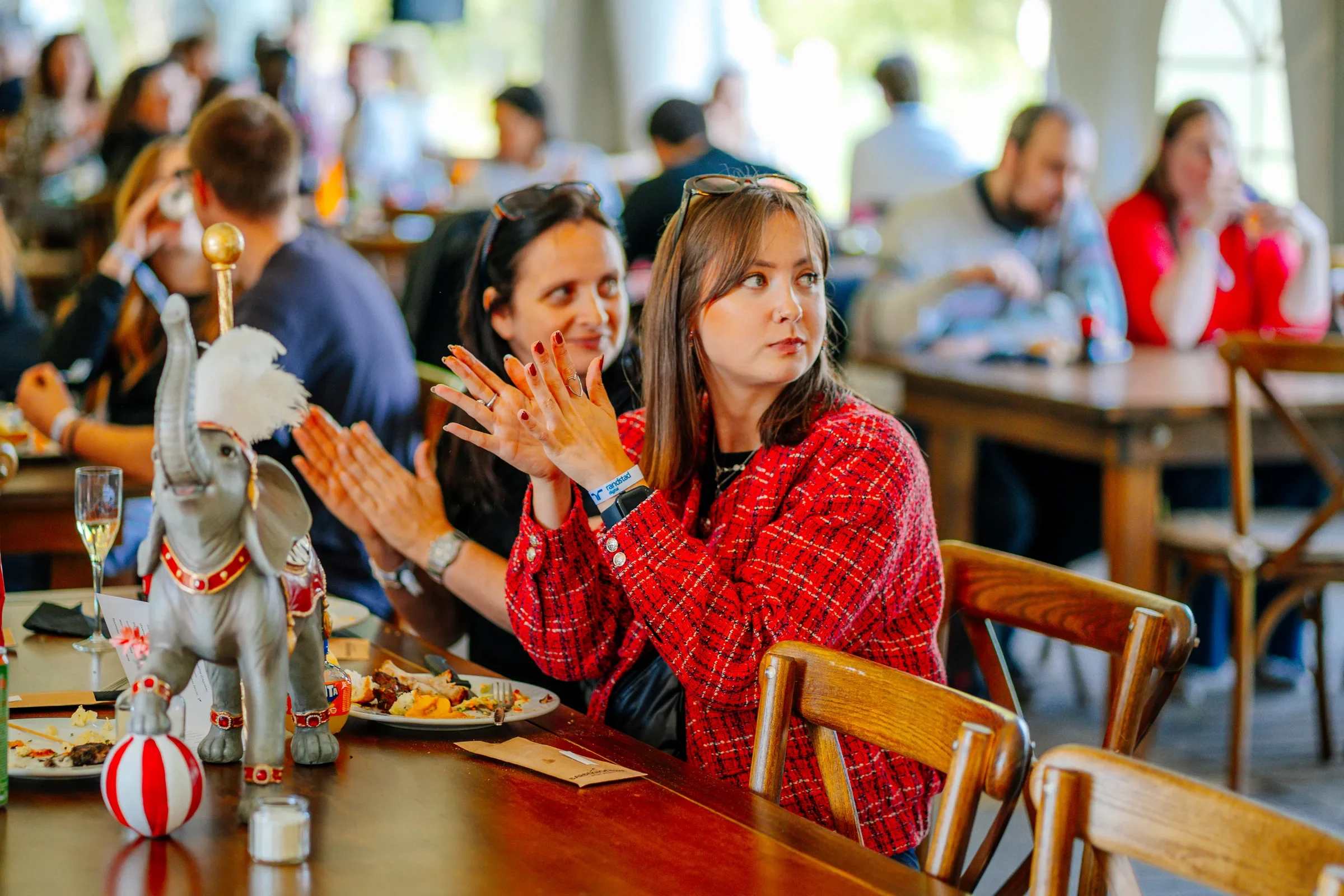 Vrouw met bruin haar en rode jas die applaudisseert tijdens een evenement met veel mensen in een grote ruimte. Op de tafel staan borden met restjes eten en decoraties van een circusachtige olifant en ballonnen.