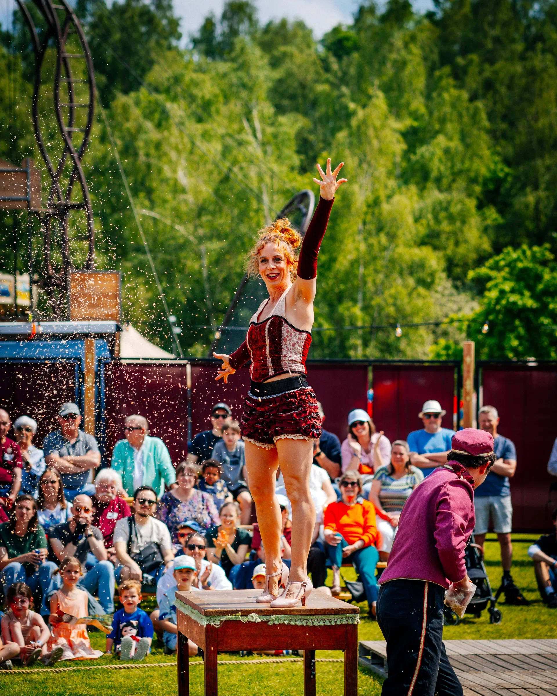 Een vrouw in een circusoutfit staat op een houten standaard en voert een show uit voor een publiek, dat in de achtergrond zit en kijkt. Het is een zonnige dag en de omgeving heeft veel groen.