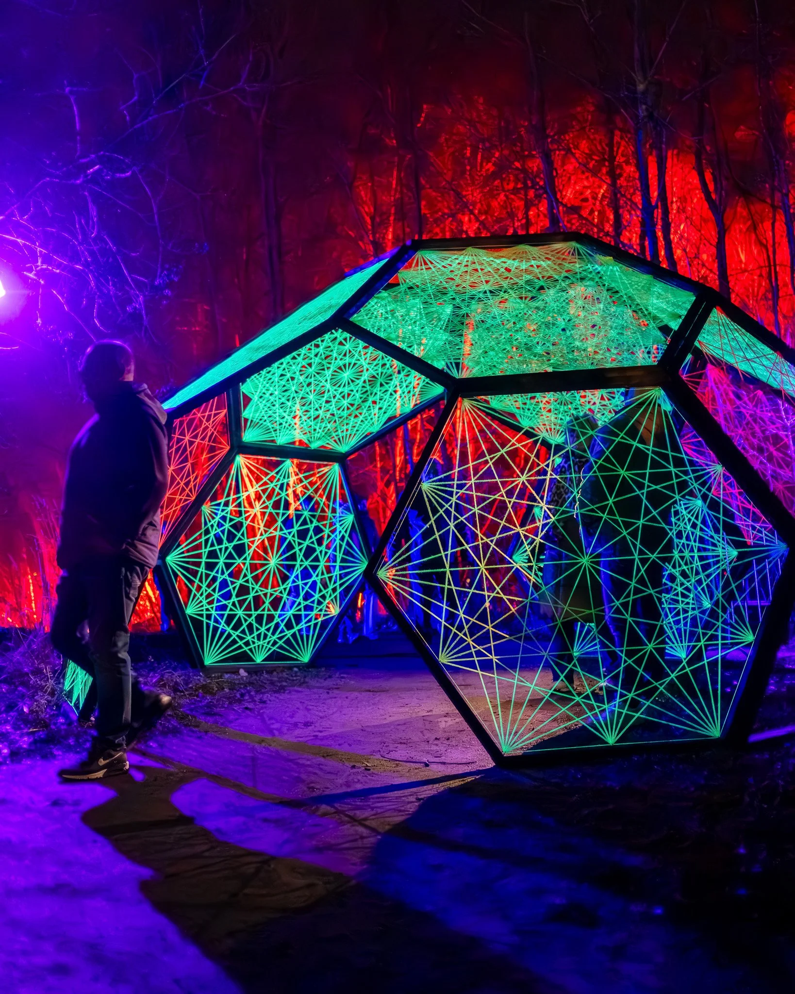 Bewolkte nacht in het bos met kleurrijke lichtinstallatie en silhouet van persoon die kijkt naar het kunstwerk.