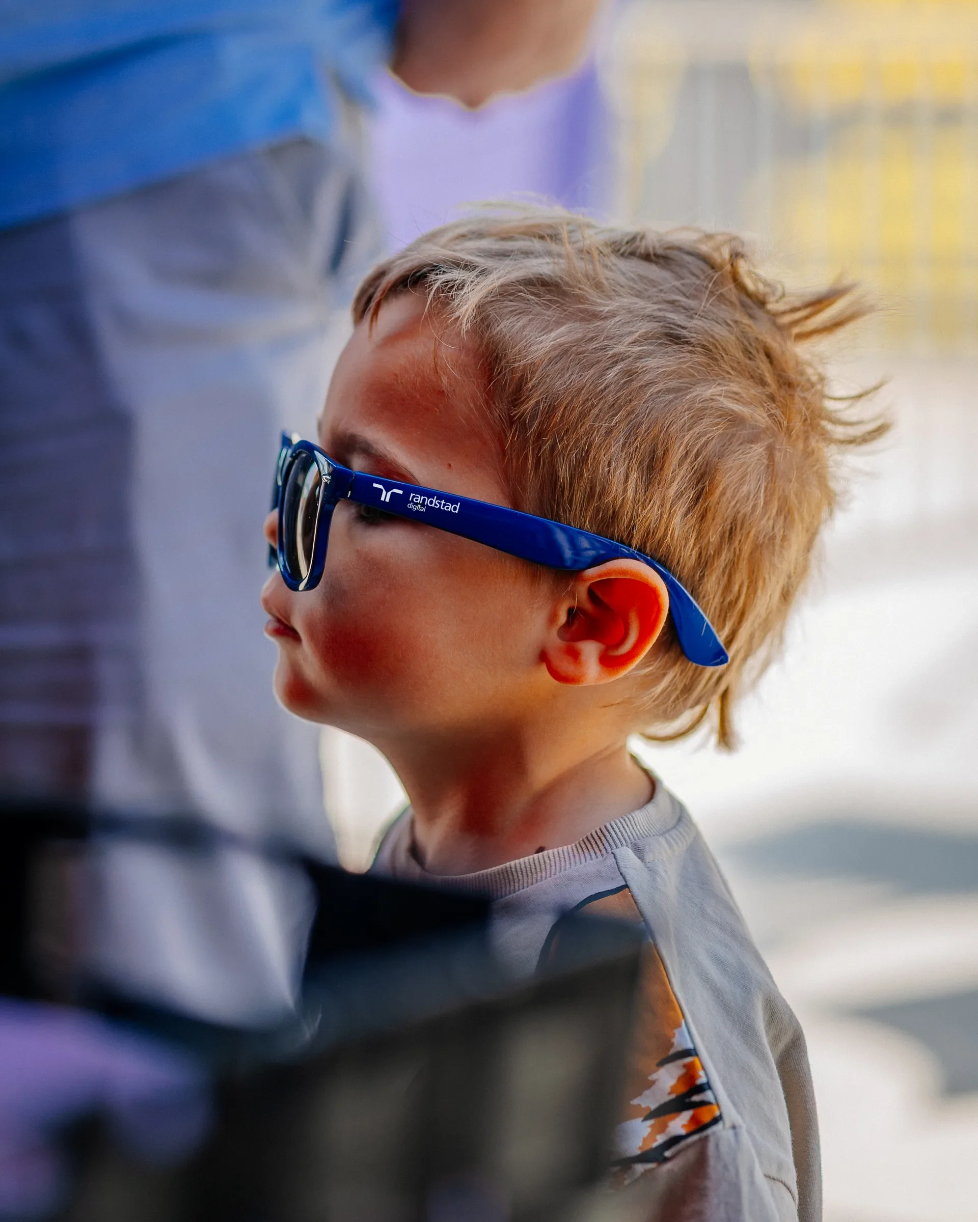 Close-up van een kind met blond haar en blauwe bril, zittend of leunend. Op de achtergrond vallen mensen en een betonnen muur. Na zonsondergang of in de schemering.