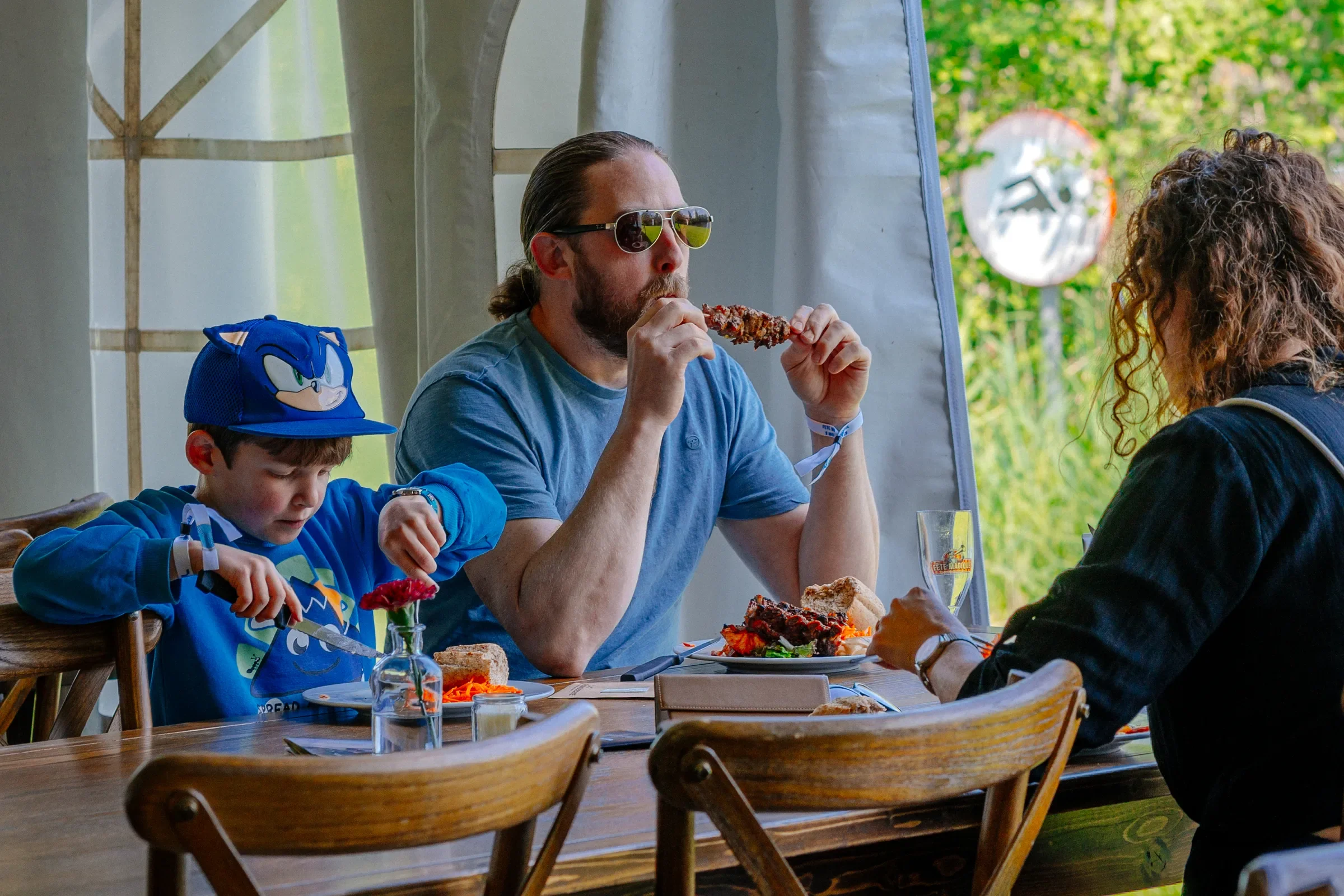 Mannen met lang haar en een baard eet vlees aan een tafel met drie andere mensen, terwijl een jongen in een blauwe sjaal en een blauwe pet naar eten kijkt. Ze zitten buiten onder een witte tent met groene bomen op de achtergrond.