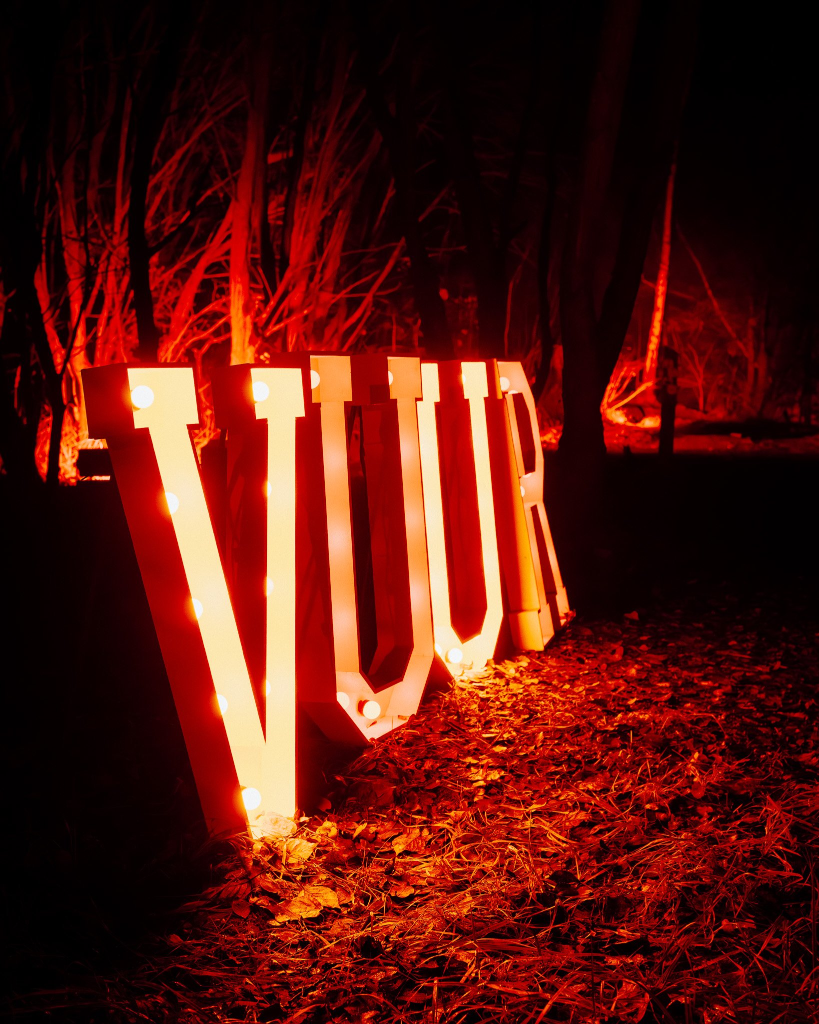 Grote verlichte letters 'LOVE' in een donkere bosrijke omgeving met rode verlichting, bladeren op de grond.