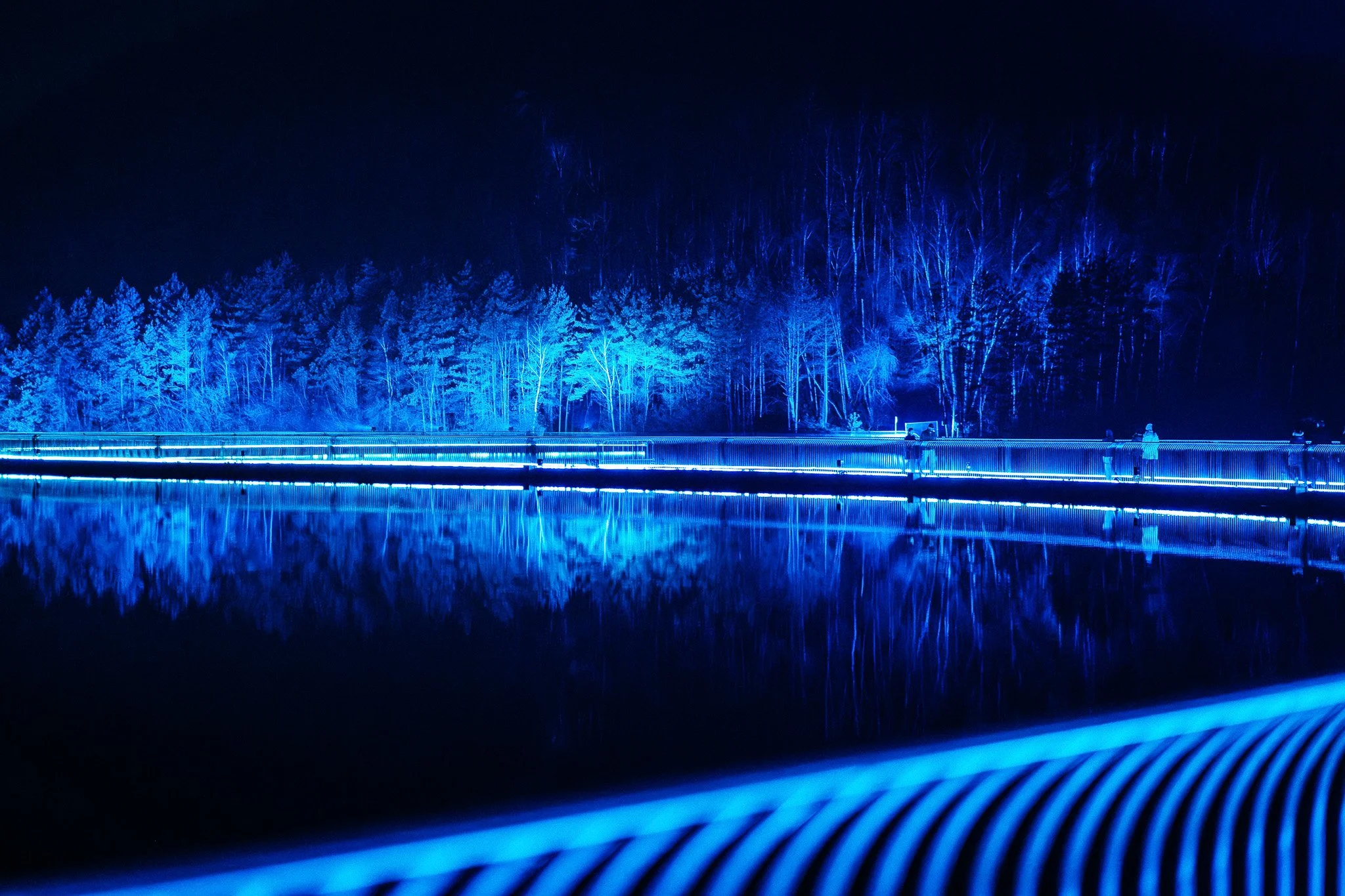 Een ​​donkerblauwe verlichte brug over een meer met een achtergrond van met sneeuw bedekte bomen en een heldere nachtelijke hemel.