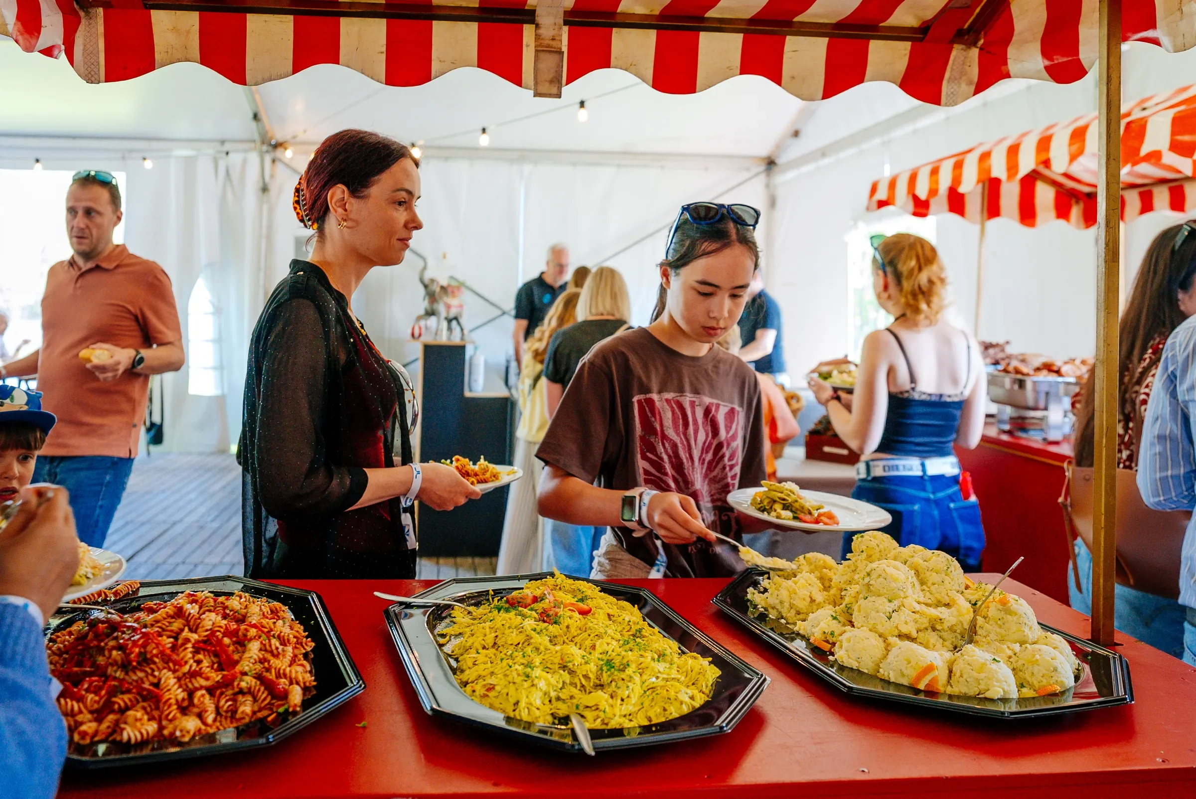 Mensen bij een buffet met verschillende gerechten, waaronder pasta, aardappelpuree en groenten, onder een tent met rode en witte strepen.