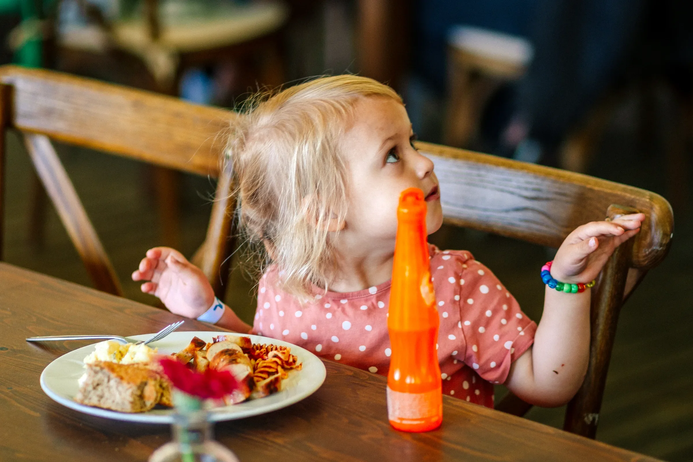 Een jonge meisje met blond haar zit aan een tafel met een bord gevuld met eten, mogelijk pizza en taart, en drinkt uit een oranje fles, in een restaurant of eetkamer.