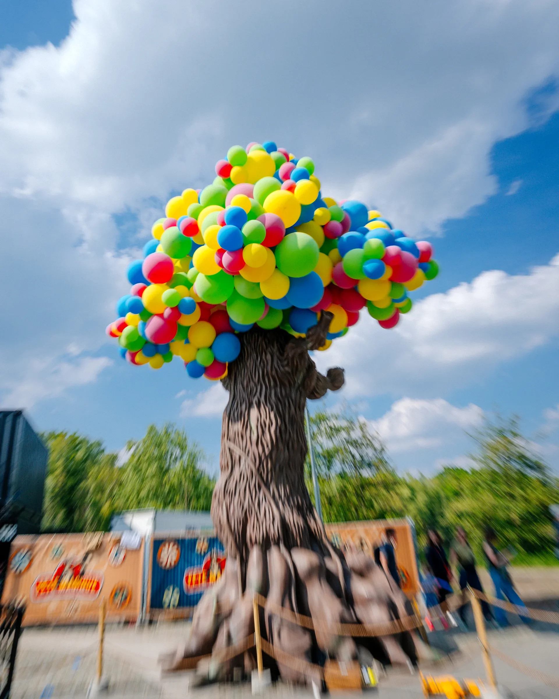 Een boom met een stam die eruitziet als een boom en een blad aan de onderkant die eruitziet als een boom, bedekt met clusters van kleurrijke ballonnen, in een buitenomgeving met een blauwe lucht en wolken.