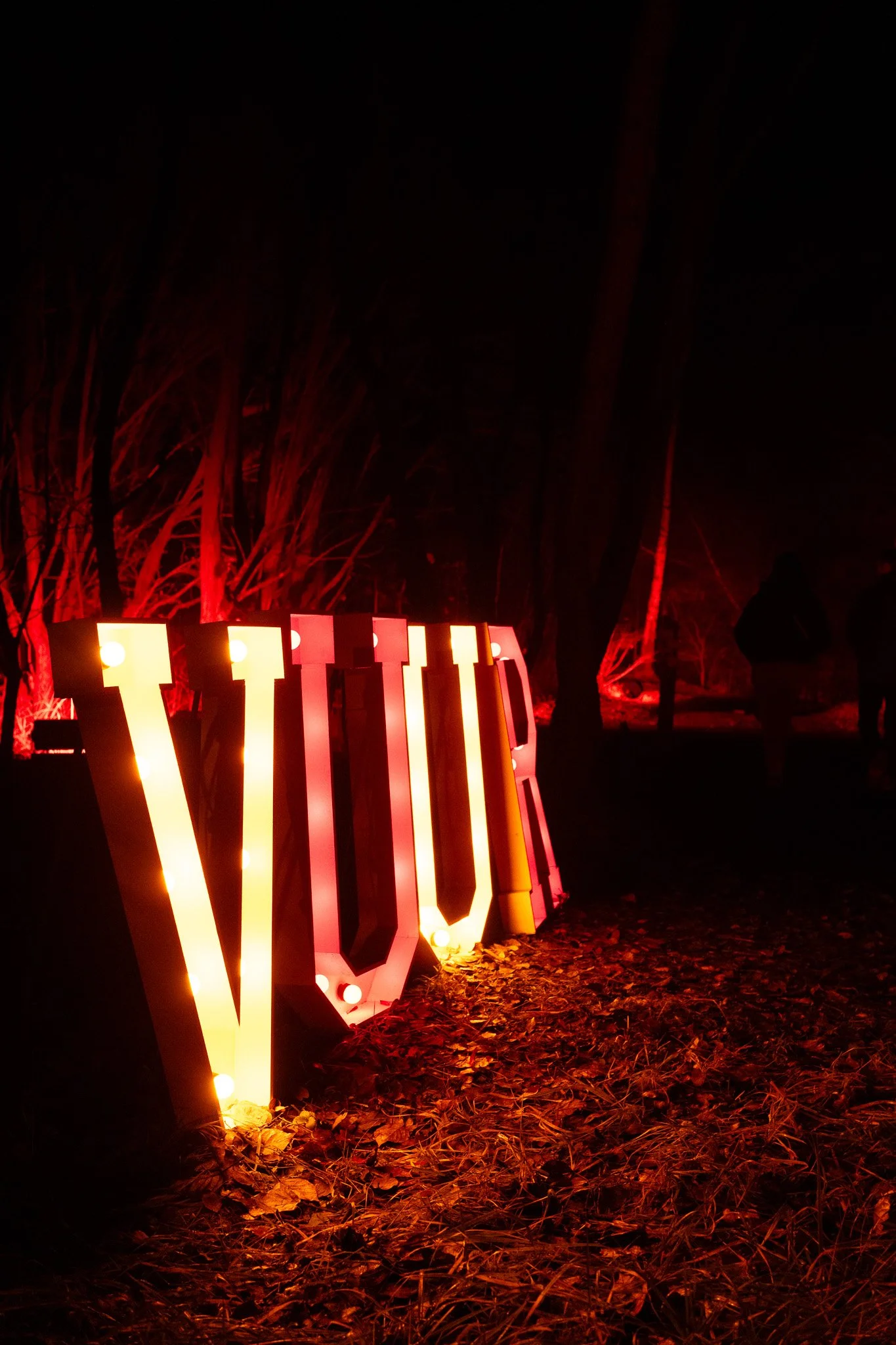 Grote, verlichte letters die samen het woord 'LOVE' vormen, geplaatst op de grond buiten, met rode verlichting op bomen op de achtergrond en een silhouet van een persoon die naar de letters kijkt.