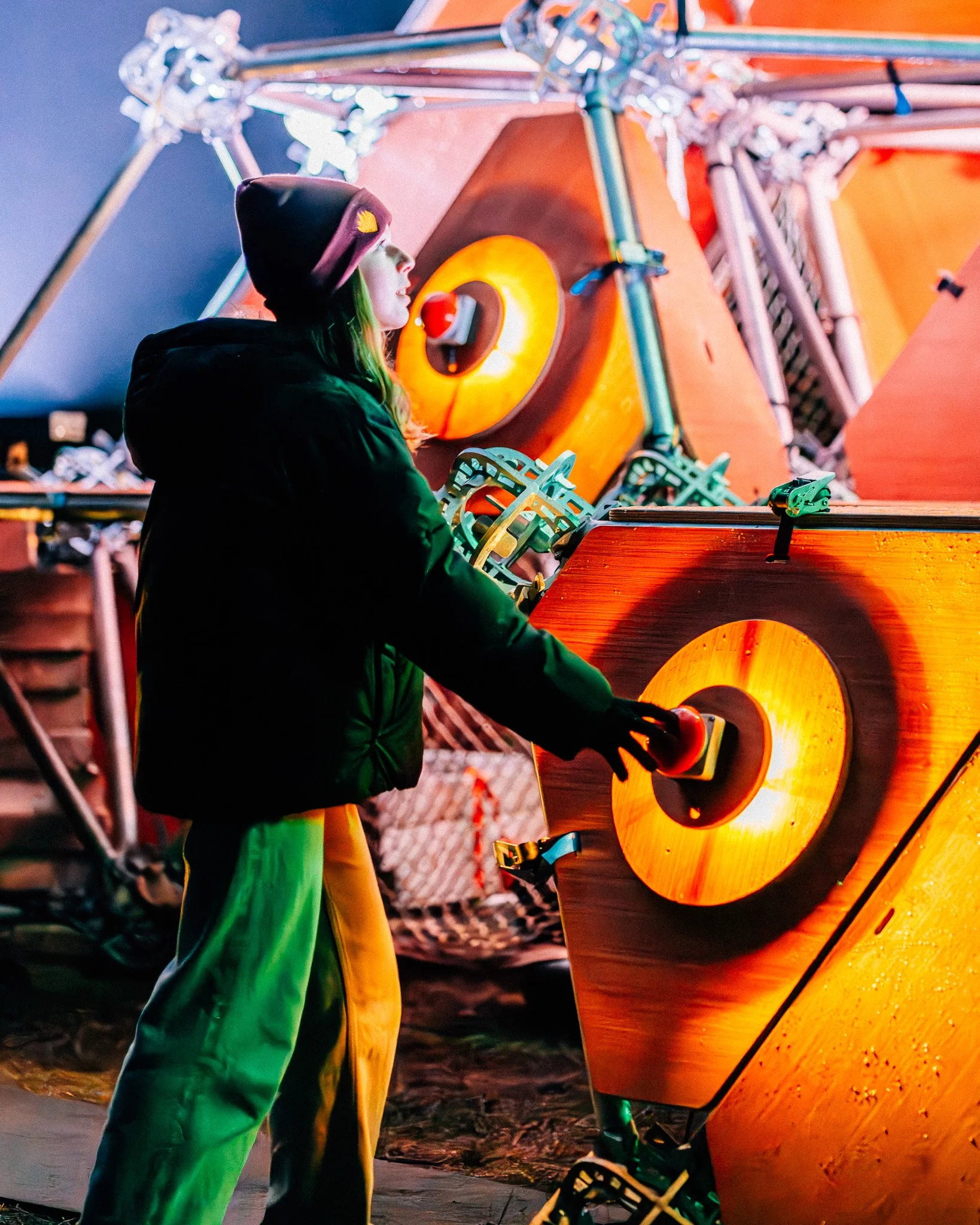 Een persoon met een donkere jas en een donker paarse muts die op een houten muziekinstrument speelt, dat lijkt op een grote, verlichte hamertje, omringd door kleurrijke metalen constructies.