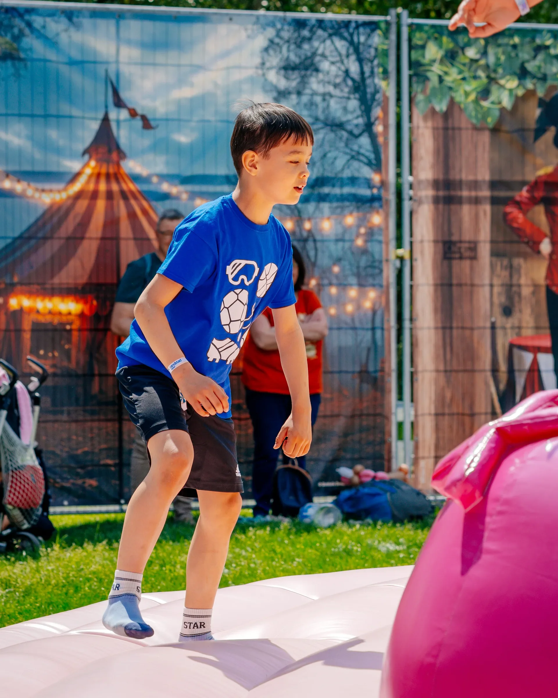 Een jongen in een blauwe T-shirt speelt op een springkussen buiten, terwijl volwassenen op de achtergrond kijken.