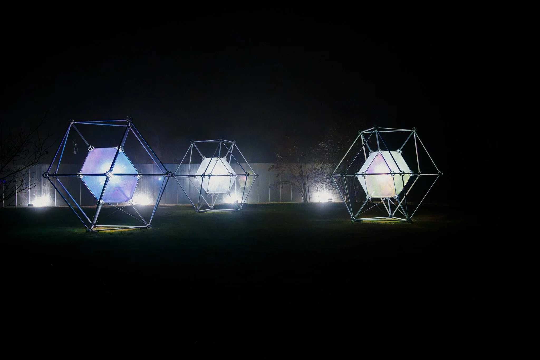 Drie geometrische lichtinstallaties in de buitenlucht 's nachts, die oplichten met neonkleuren, met donkere hemel en enkele bomen op de achtergrond.