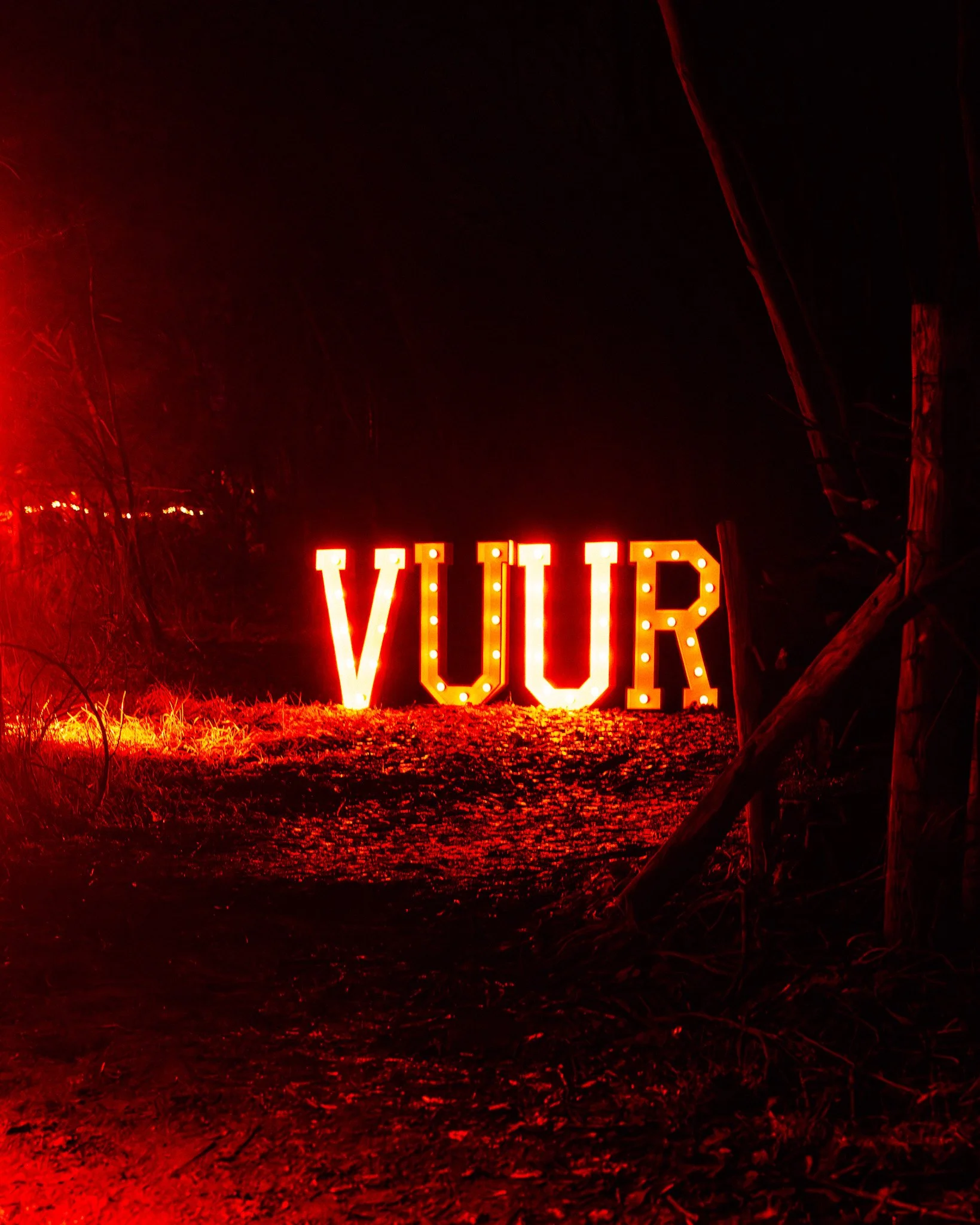 Verlichte letters die het woord 'VUI' vormen in een duistere omgeving, omgeven door bomen en struiken, met een rode gloed en een mistige sfeer.