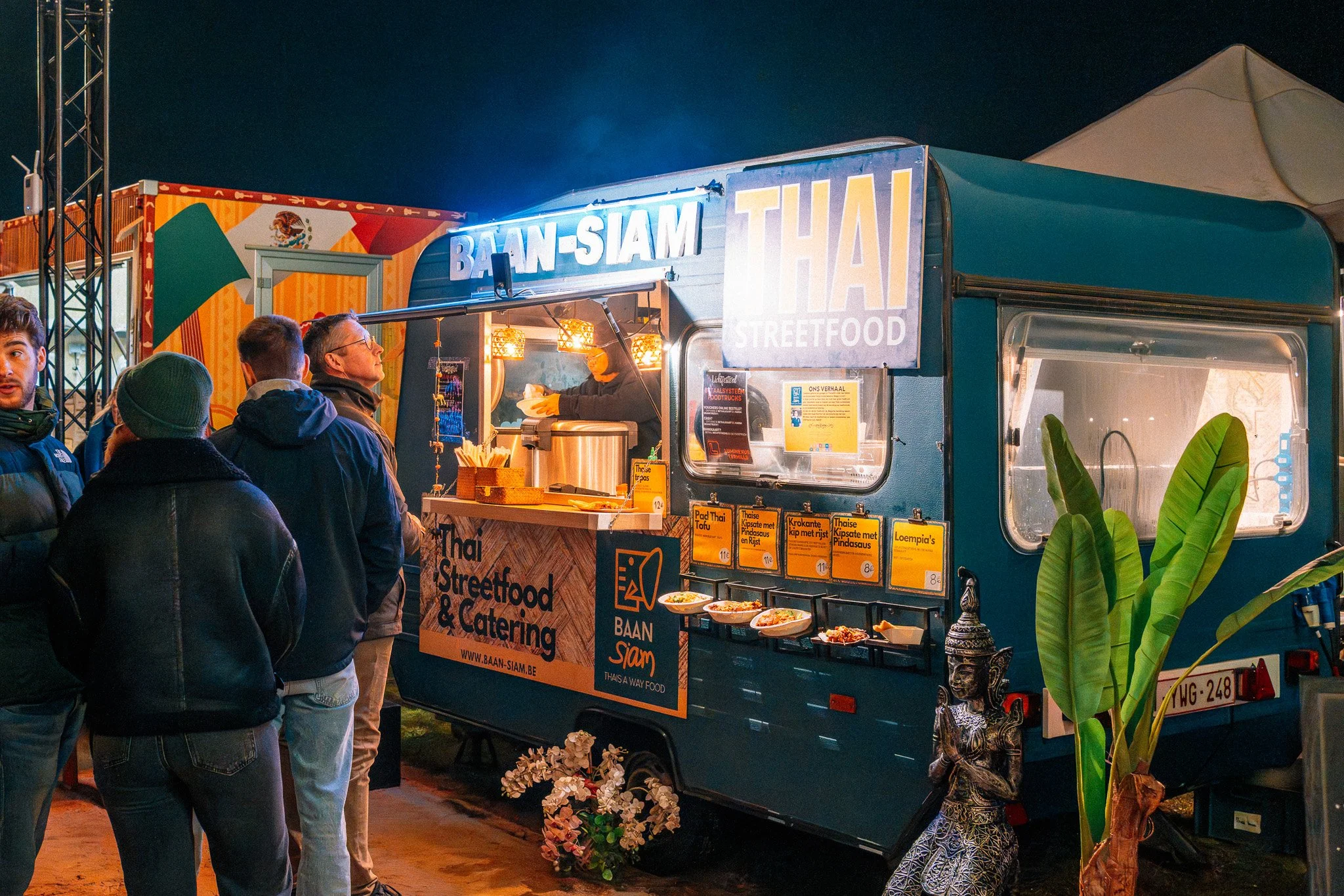 Een foodtruck verkoopt streetfood aan mensen 's avonds, met een menu op de muur en versierd met planten en bloemen.