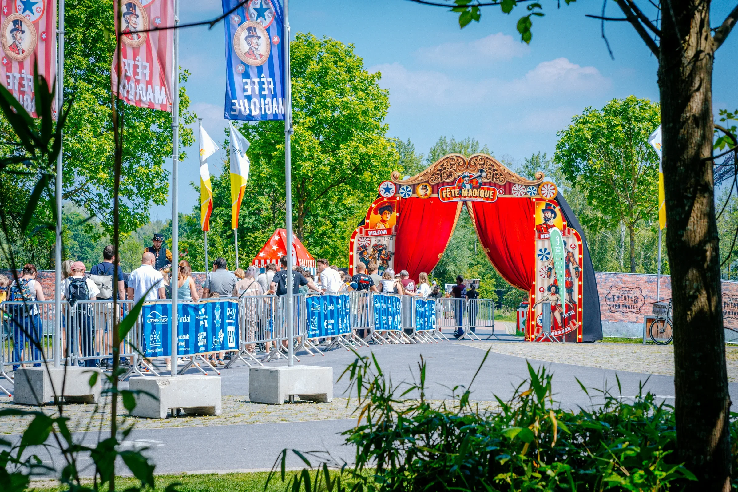 Bezoekers wachten in een rij voor een circustheater met rode gordijnen, omringd door groene bomen, vlaggen en een blauwe lucht.