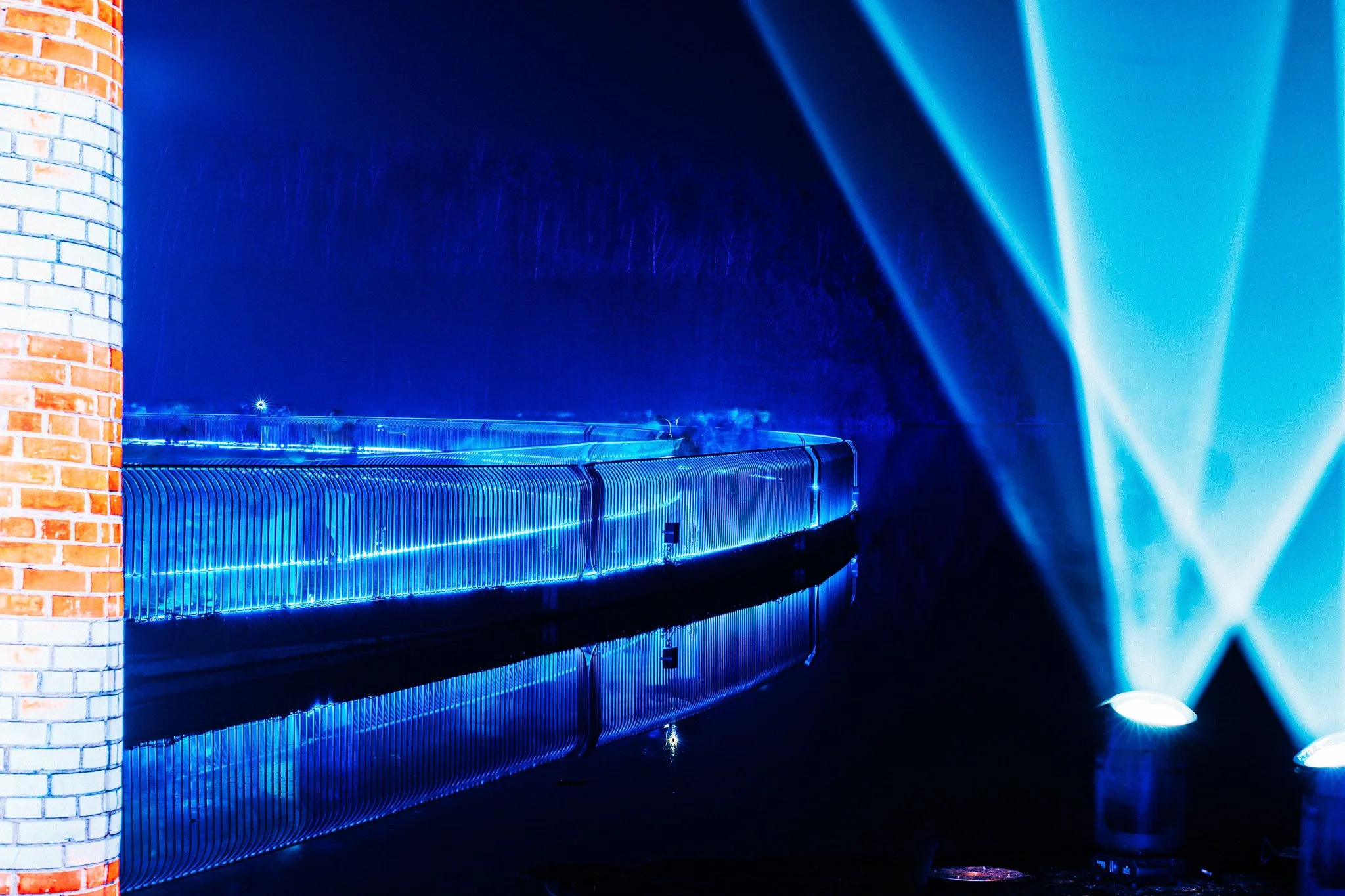 Een brug met blauwe LED-verlichting en een bakstenen muur aan de linkerzijde, nachtelijke setting, met blauwe lichtstralen in de rechterbovenhoek.
