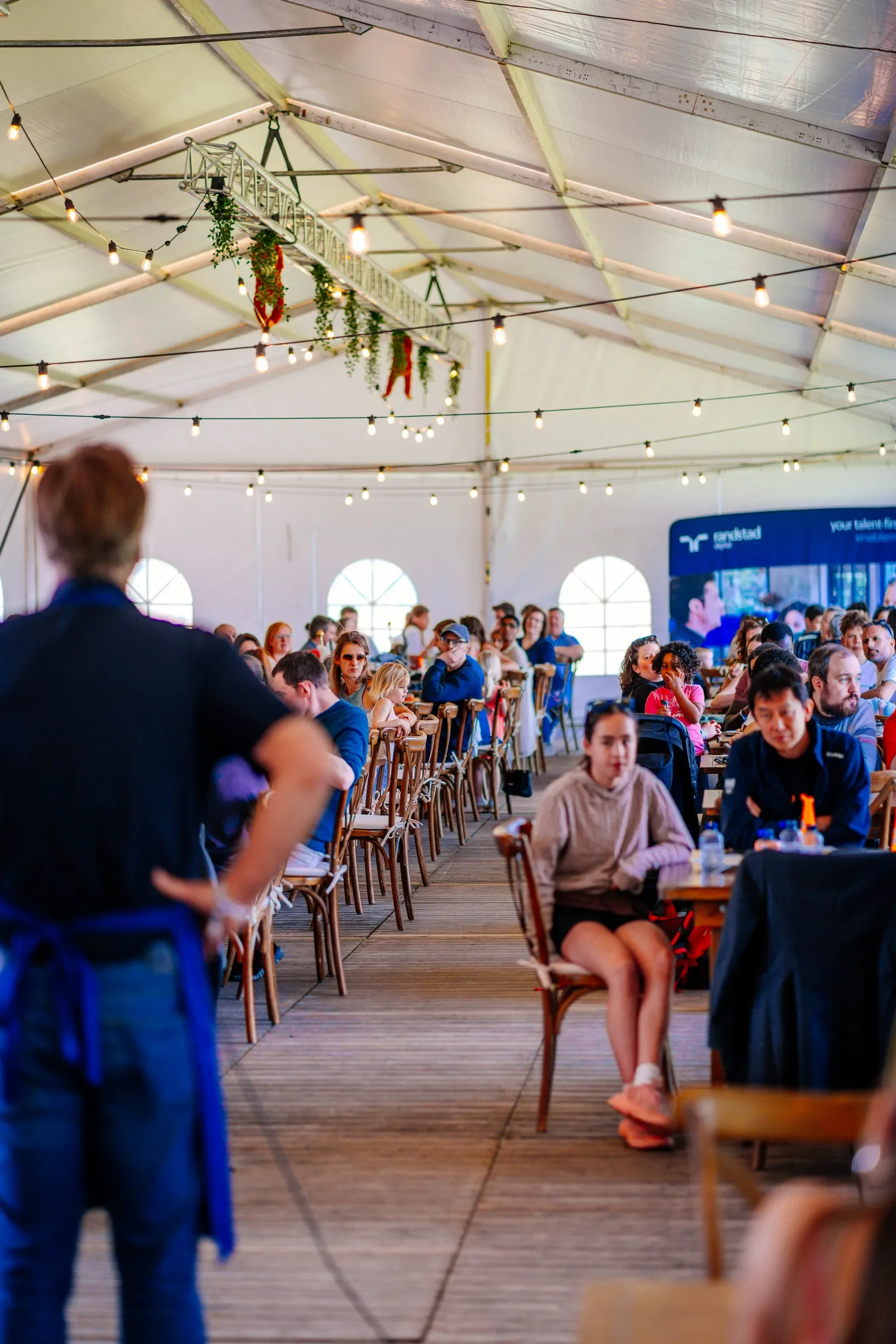 Binnen in een grote tent dat wordt gebruikt voor een evenement of een catering, met veel gasten die aan tafels zitten. De tent is versierd met stringverlichting en een decoratief element aan het plafond.