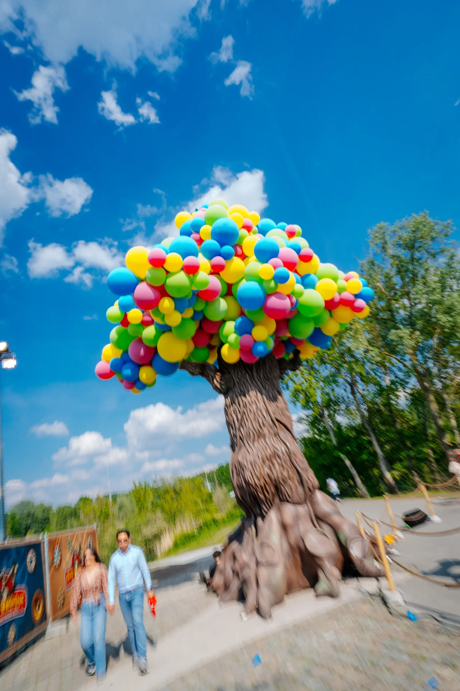 Een sculptuur van een boom met een stam en een bladerdek gemaakt van kleurrijke ballonnen in verschillende kleuren zoals geel, blauw, roze en groen, onder een blauwe hemel met wat wolken.