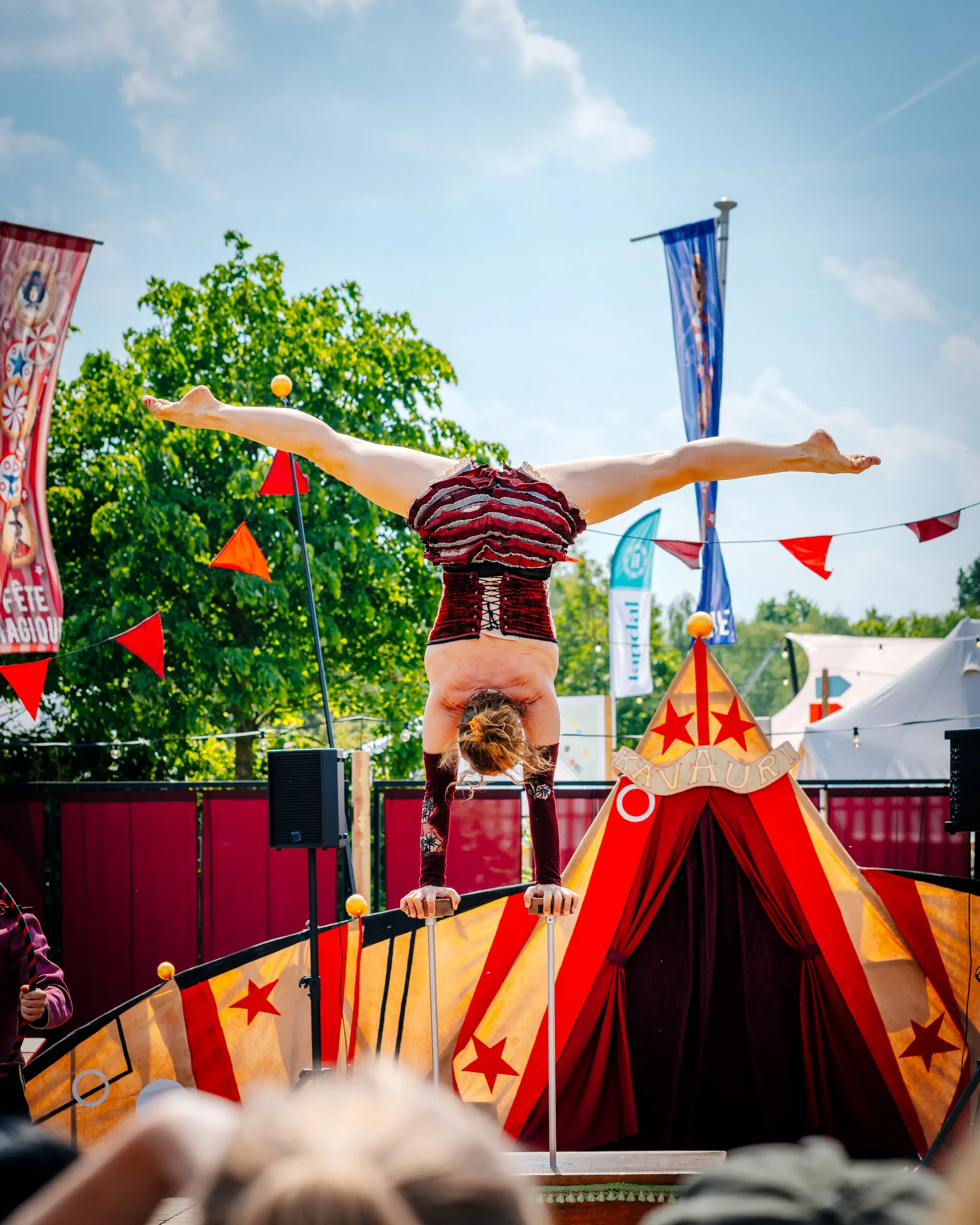 Een acrobaat die een handstand doet op een circuspodium, met armen gestrekt en benen gespreid, onder een grote rode en gele circustent, omringd door vlaggen en een kleurrijke achtergrond met bomen en een blauwe lucht.