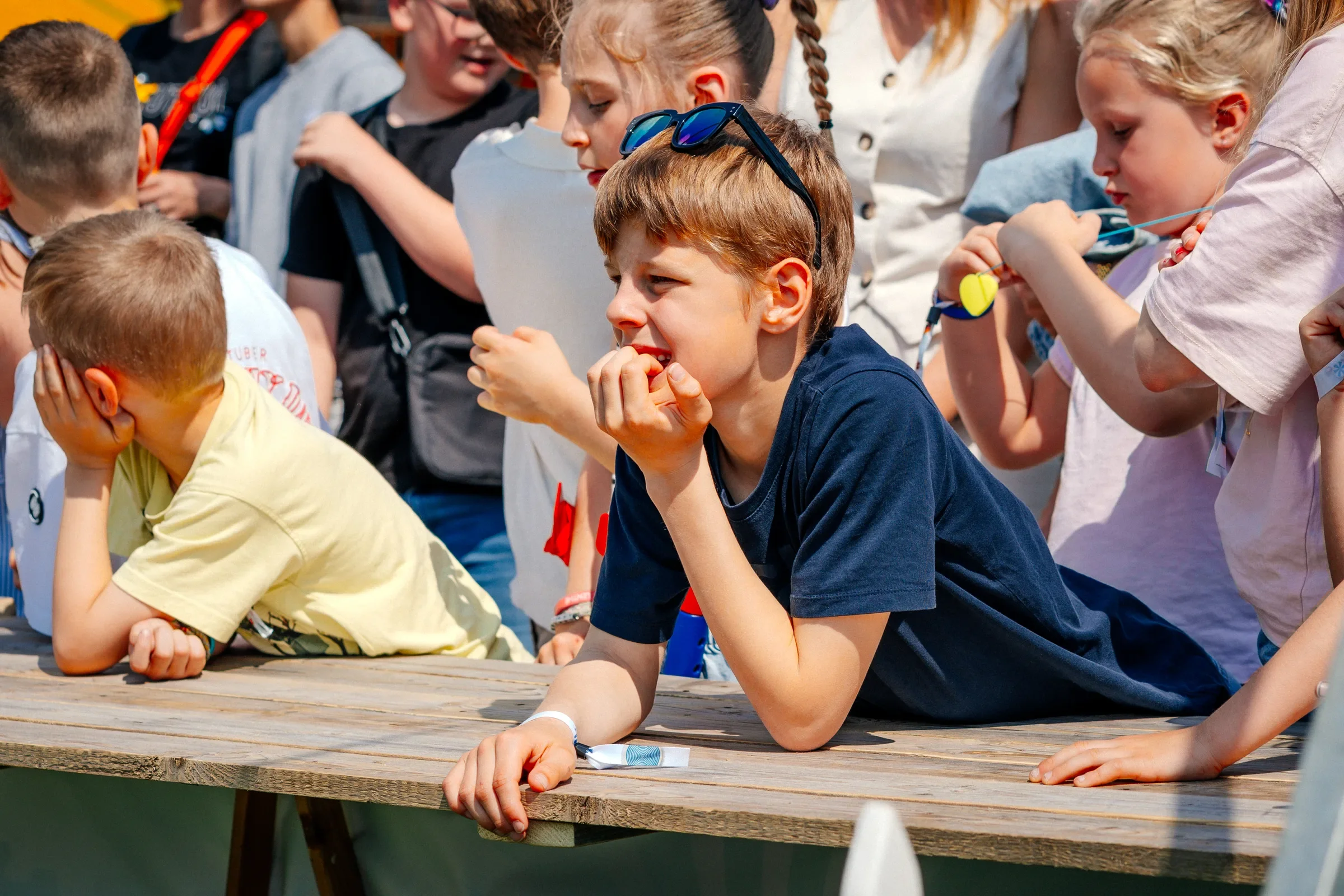 Kinderen zitten op een houten tafel buiten, sommige lijken na te denken of te wachten, andere kijken geconcentreerd. Ze dragen casual kleding en hebben accessoires zoals zonnebrillen en armbanden.