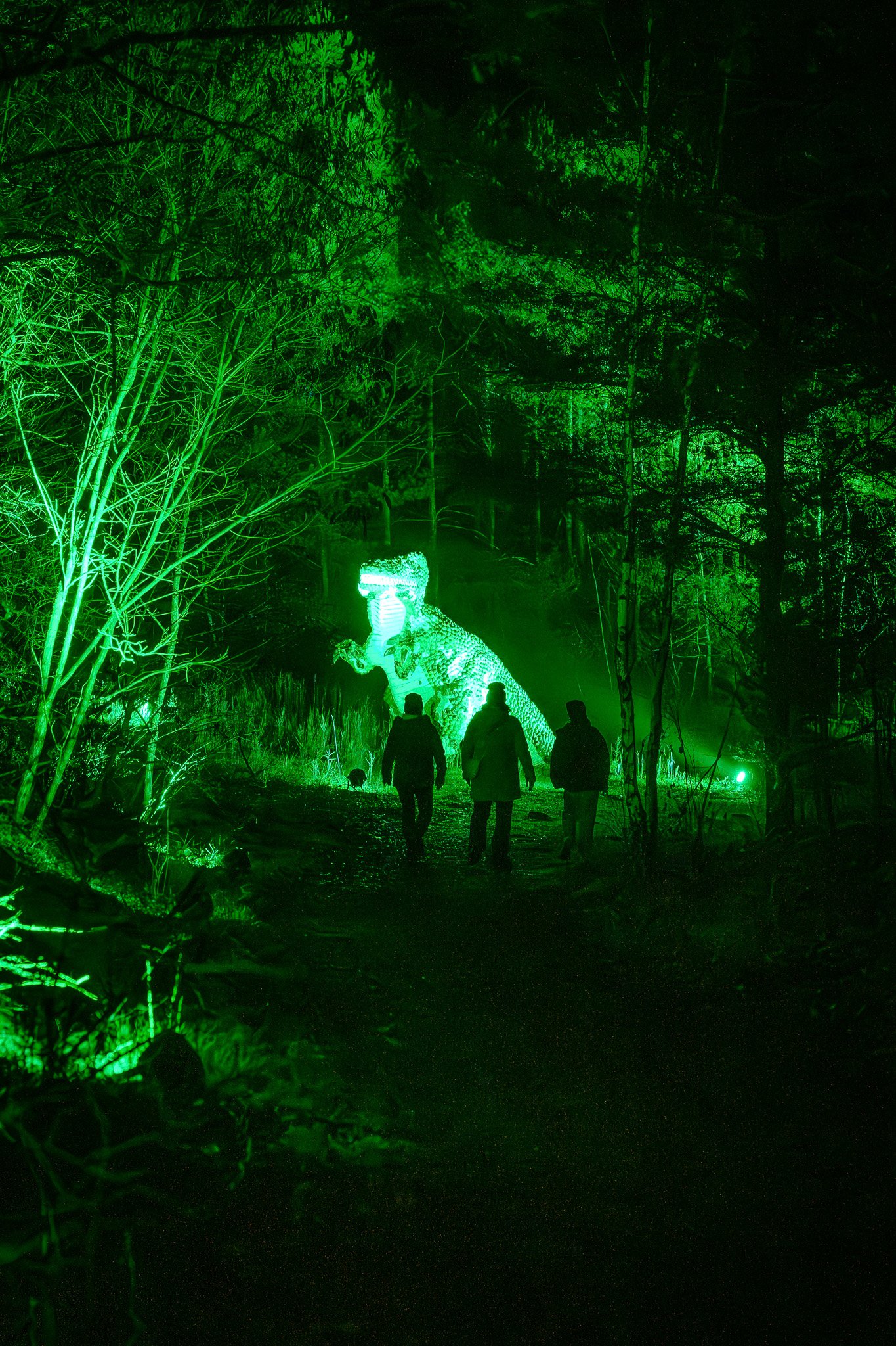 Mensen wandelen door een bos met groene verlichting en een verlichte dinosaurus figuur op de achtergrond.