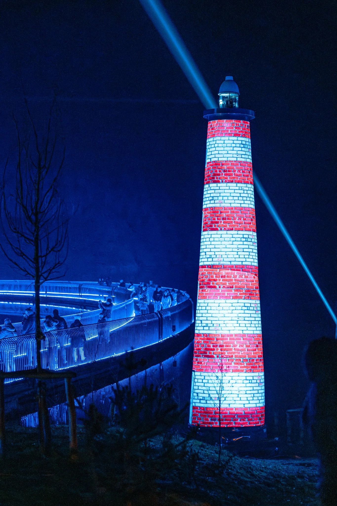 Een verlichte vuurtoren met rode en witte strepen in de nacht, met een lichtstraal die de lucht in schijnt. Er is een donkere diepte onderaan en een treinrails met een groep mensen erlangs, die in de blauwe schijn van licht staan.