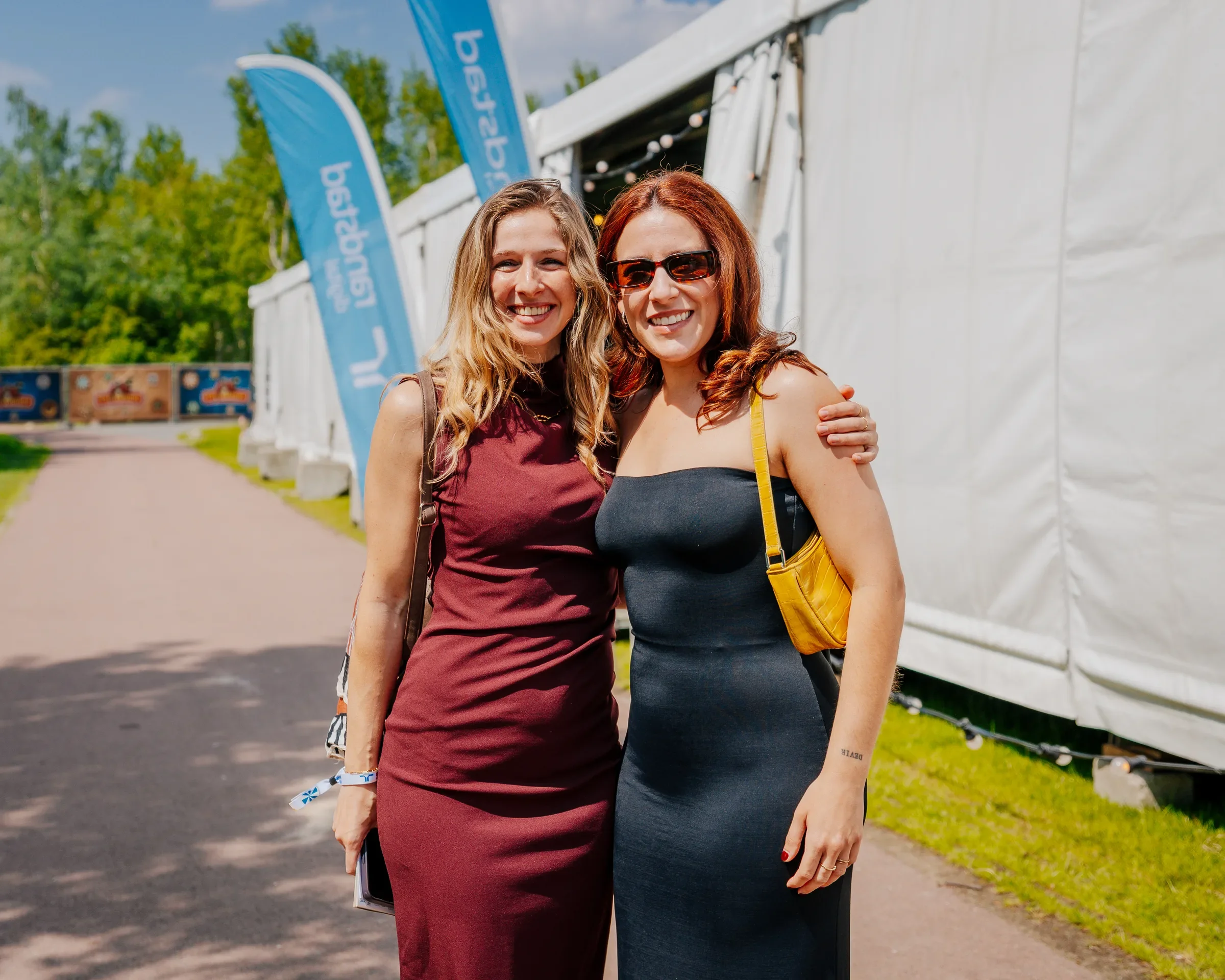 Twee vrouwen staan dicht bij elkaar en lachen, met een festivalvlag en een witte tent op de achtergrond op een zonnige dag.