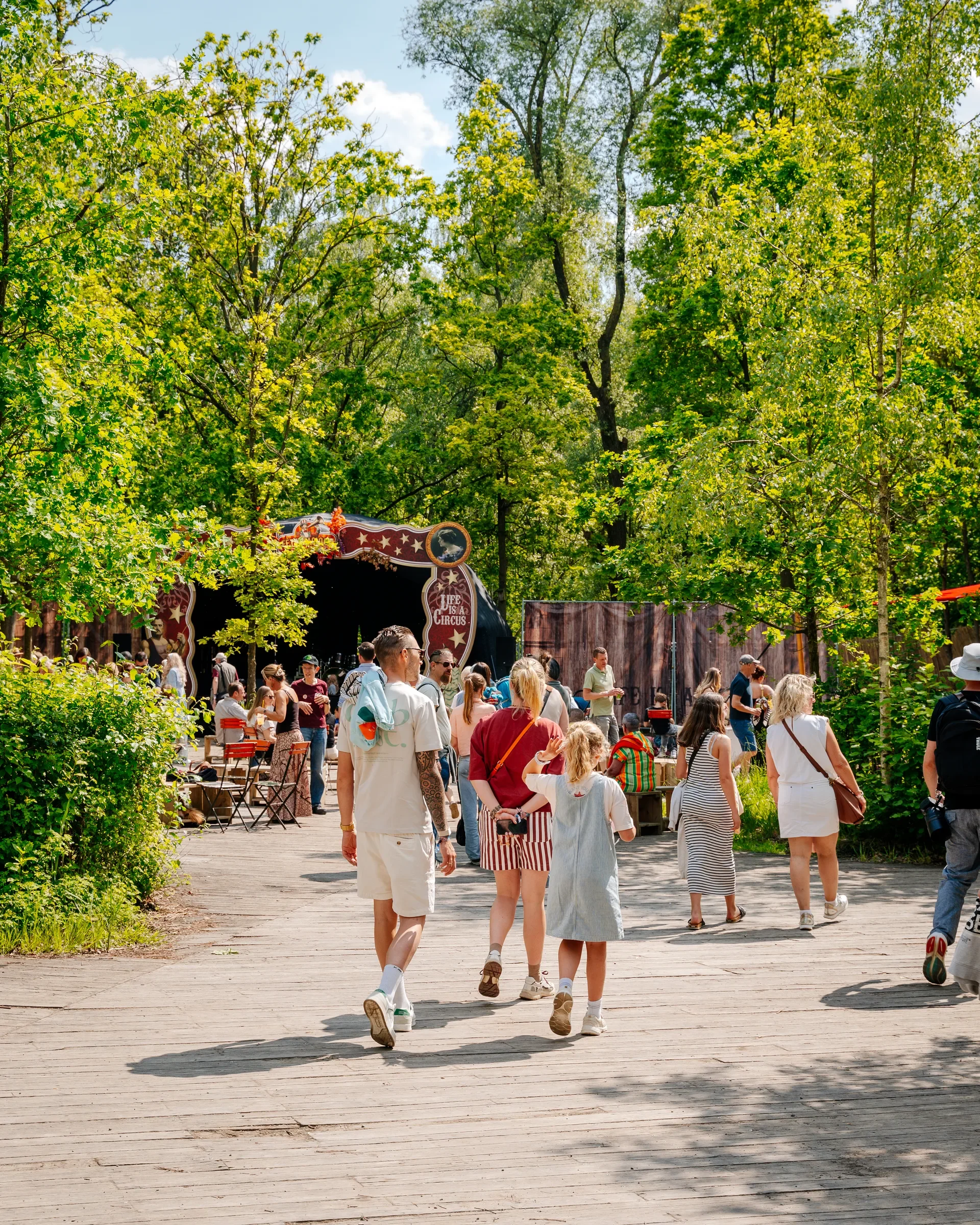 Mensen lopen over een houtenpad in een park met veel groen, bomen en een circussleur, terwijl ze genieten van de zonnige dag.