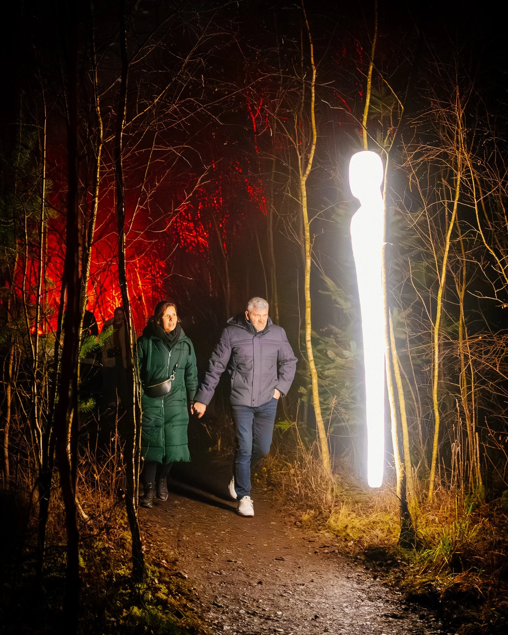 Twee mensen wandelen op een pad door een bos 's nachts, verlicht door een grote verticale lamp en rode verlichting op de achtergrond.
