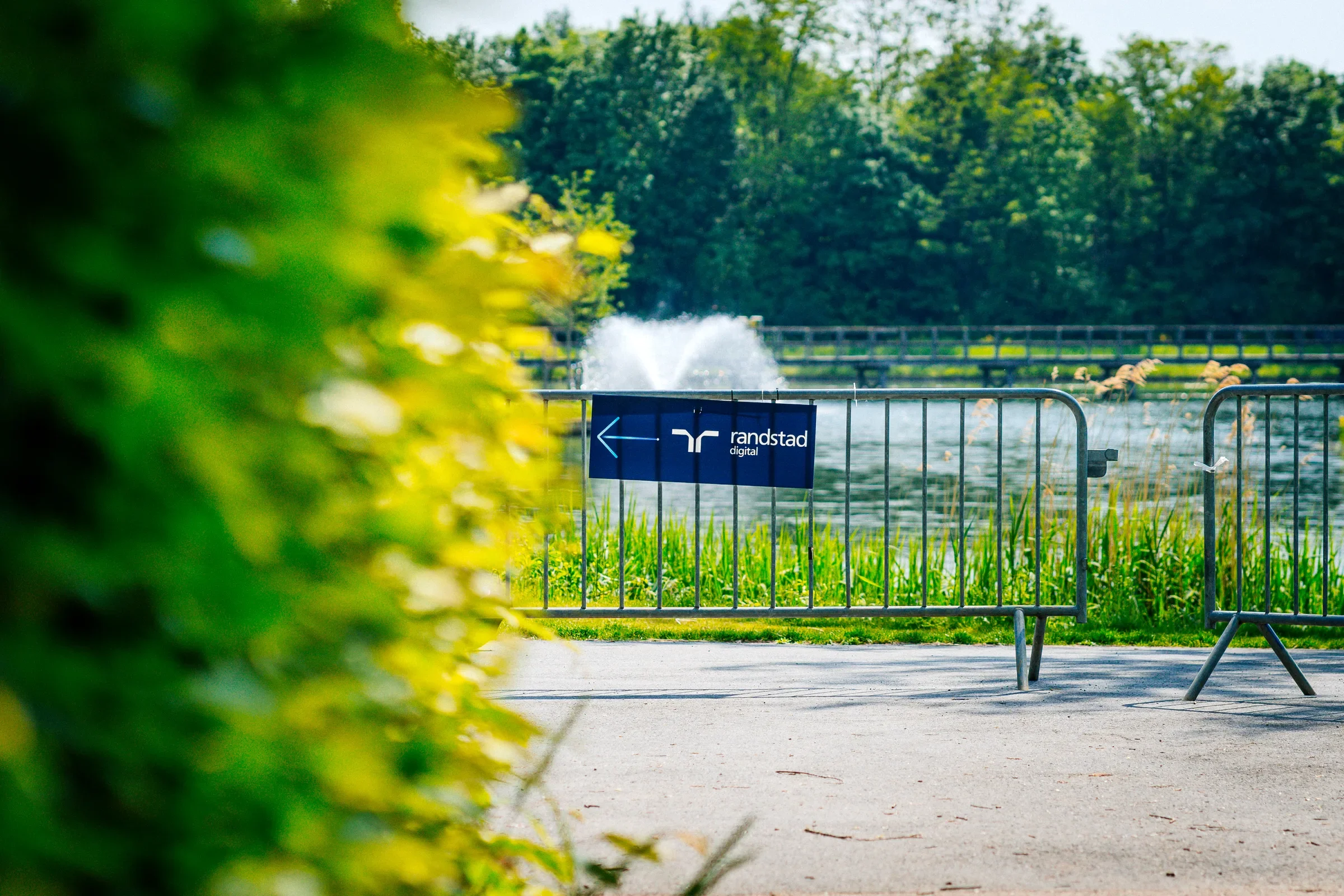 Een vaart met een waterfontein, omgeven door groene bomen en een hek met een bord dat 'Randstad Digital' advertentie bevat.
