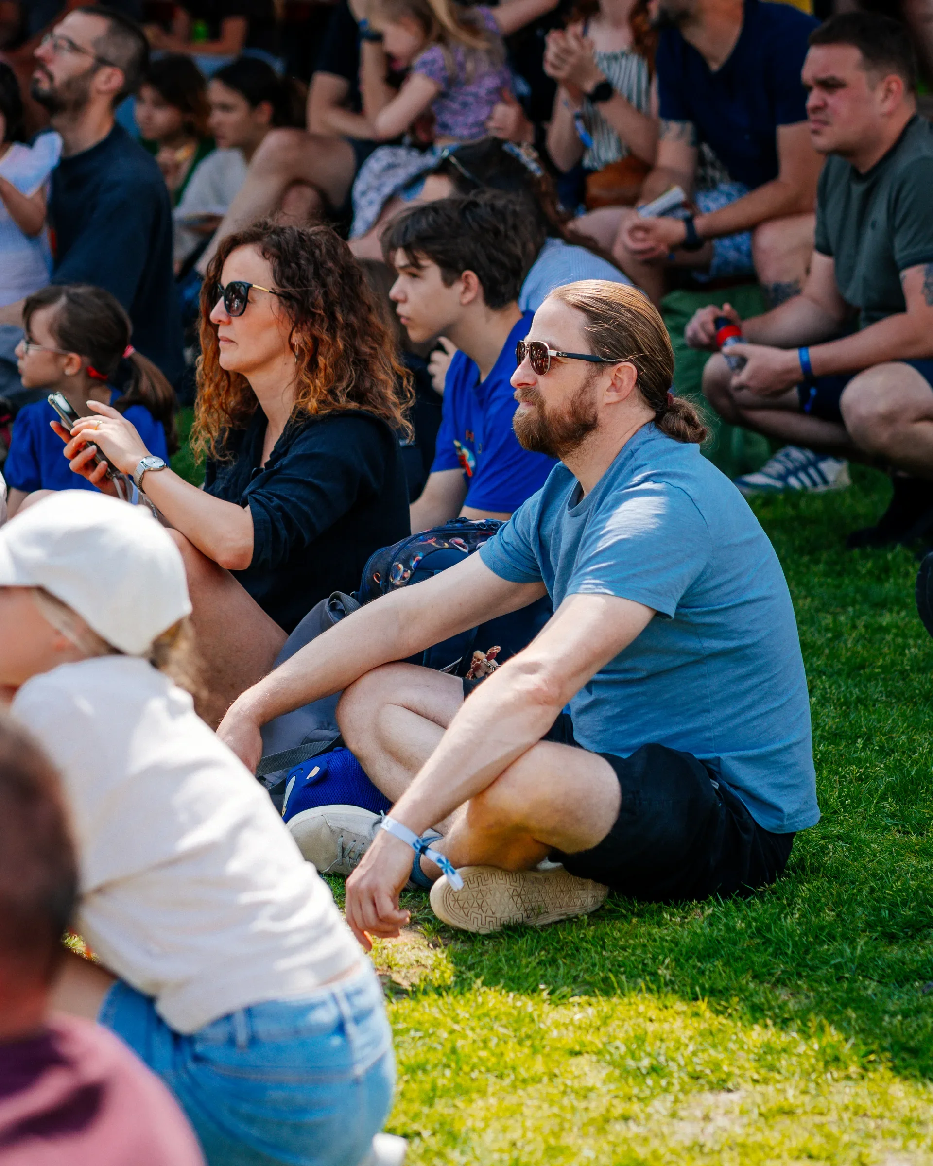 Mensen zitten op gras bij een evenement, sommige dragen zonnebrillen en zijn geconcentreerd.