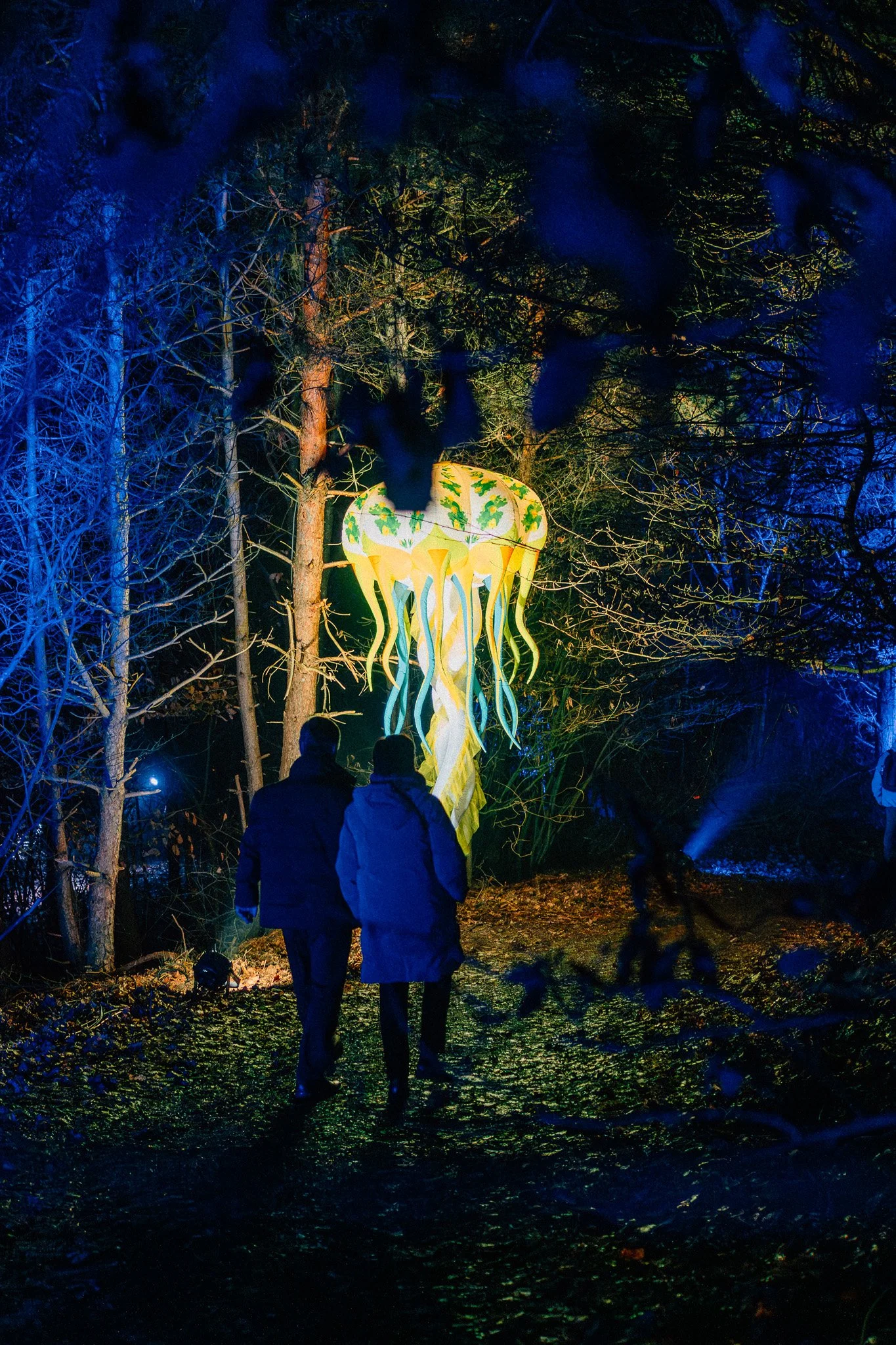 Twee mensen lopen in een donker bos met bomen, terwijl een verlichte kunstenaarsinstallatie in de achtergrond op een neerwaartse houtachtige structuur staat. De installatiestructuur is geel met groene bladeren en lange hangende elementen.