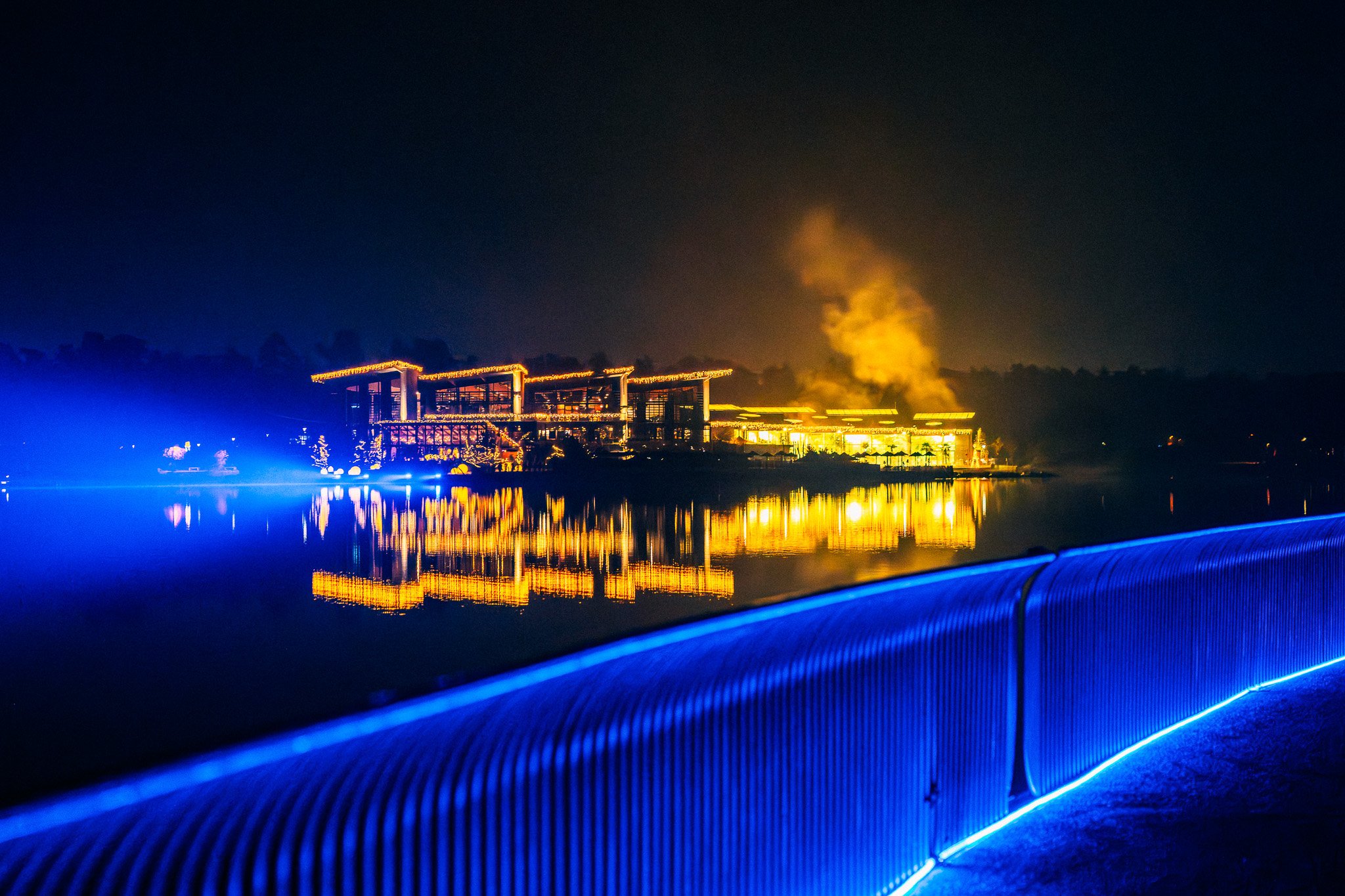 Gebouwen en een hefbrug bij nacht, verlicht door gele en blauwe lichten, met een reflectie in het water ervoor.