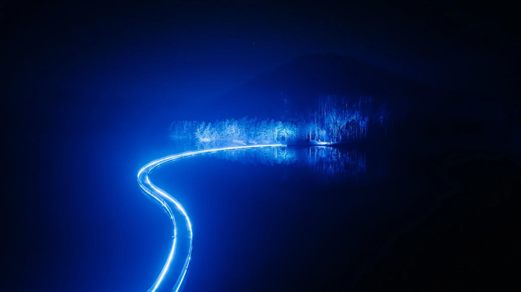 Lichtspoor dat door een rustige wateroppervlakte reflecteert, met een bos en bergen op de achtergrond, onder een nachtelijke hemel.