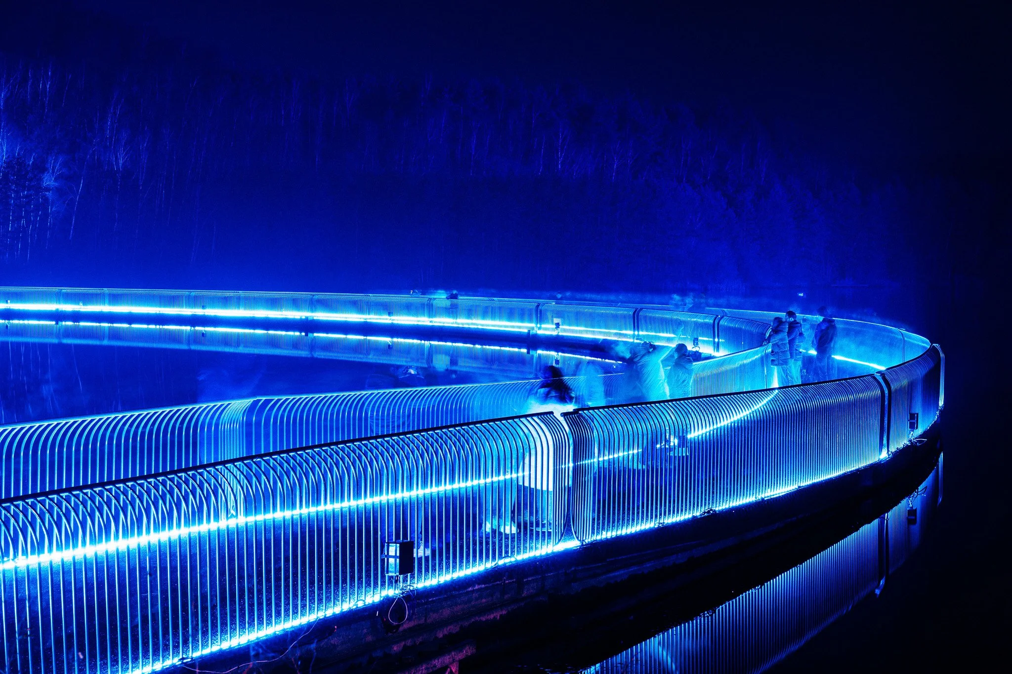Mensen staan op een wandelbrug met blauwe LED-verlichting 's nachts, omgeven door donkere bomen.