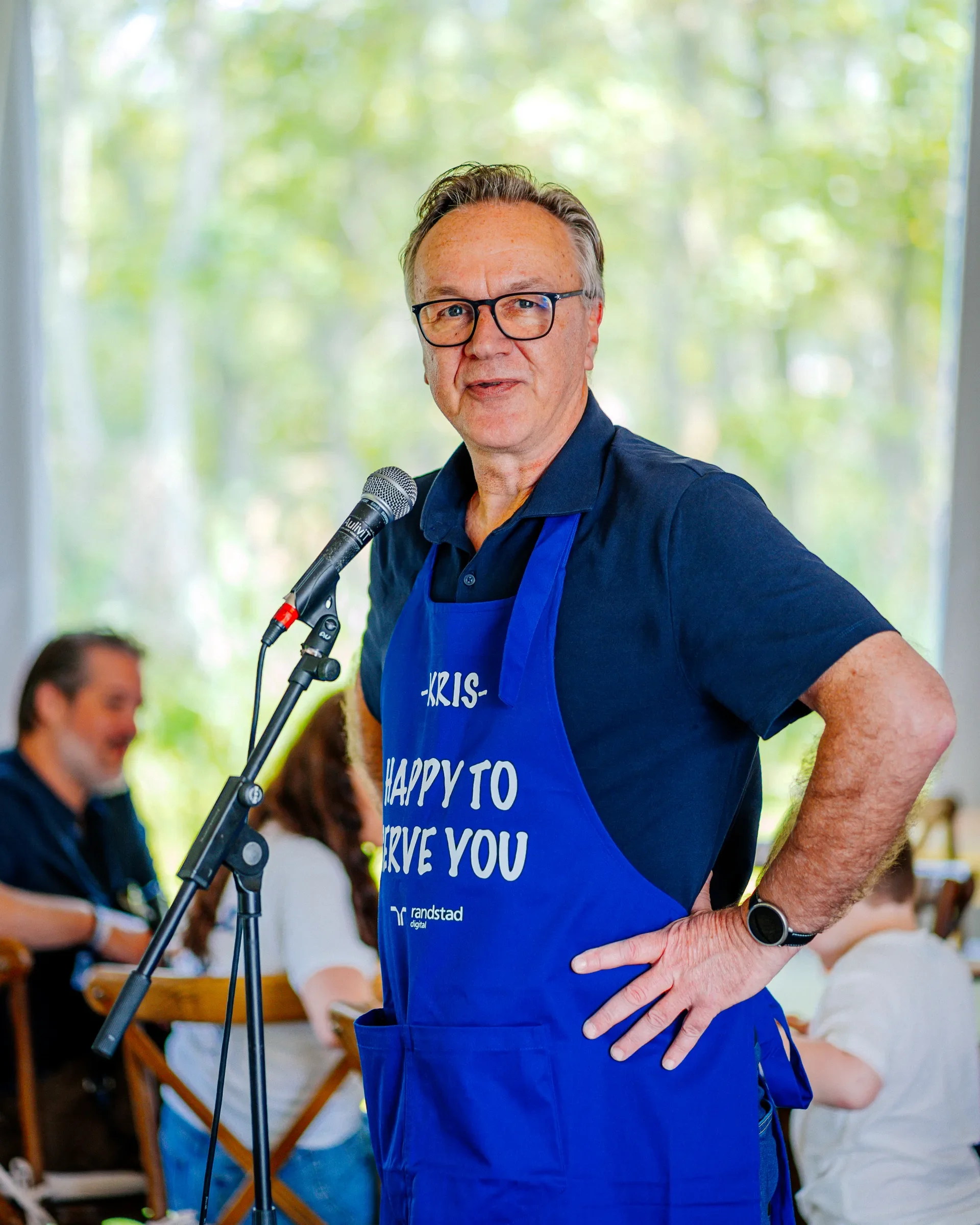 Man met bril en blauwe apron die voor een microfoon staat, met mensen en buiten in de achtergrond.