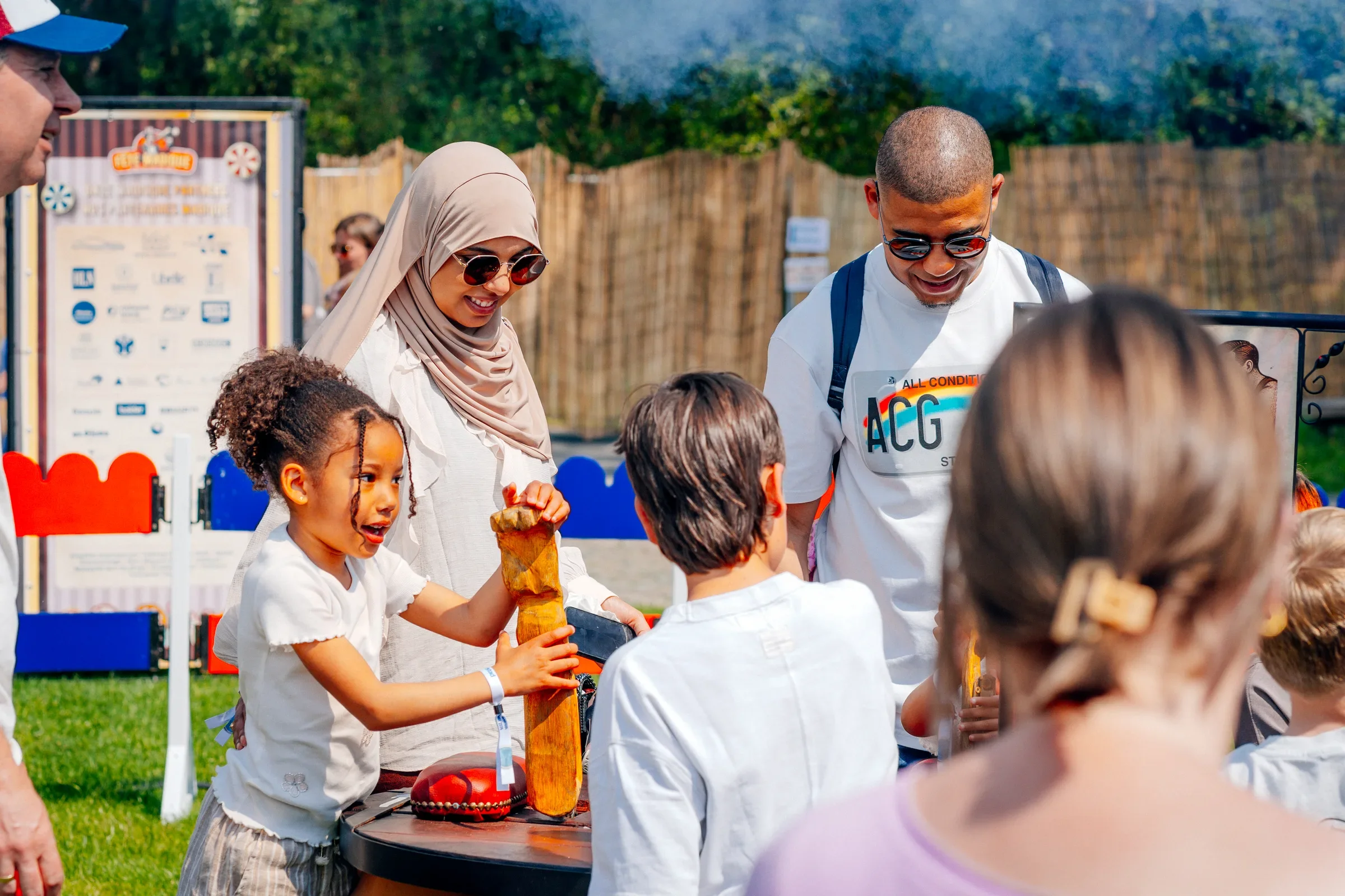 Kinder en volwassenen genieten van een activiteit buiten op een zonnige dag, met een houten fence en bomen op de achtergrond.