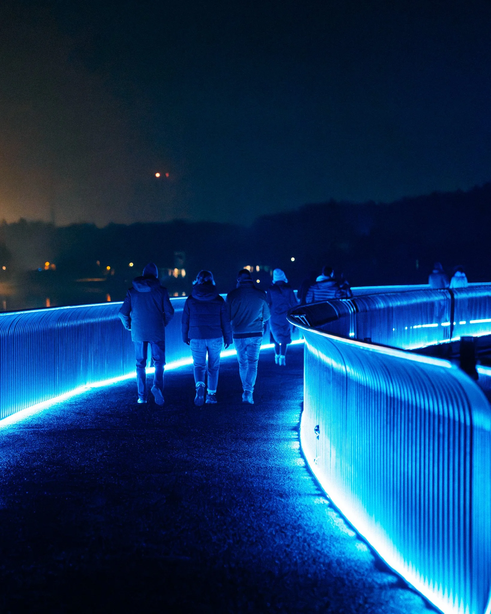 Mensen wandelen over een weg met blauwe verlichting in de avond of nacht, tegen een donkere hemel met enkele rode lichten zichtbaar op afstand.