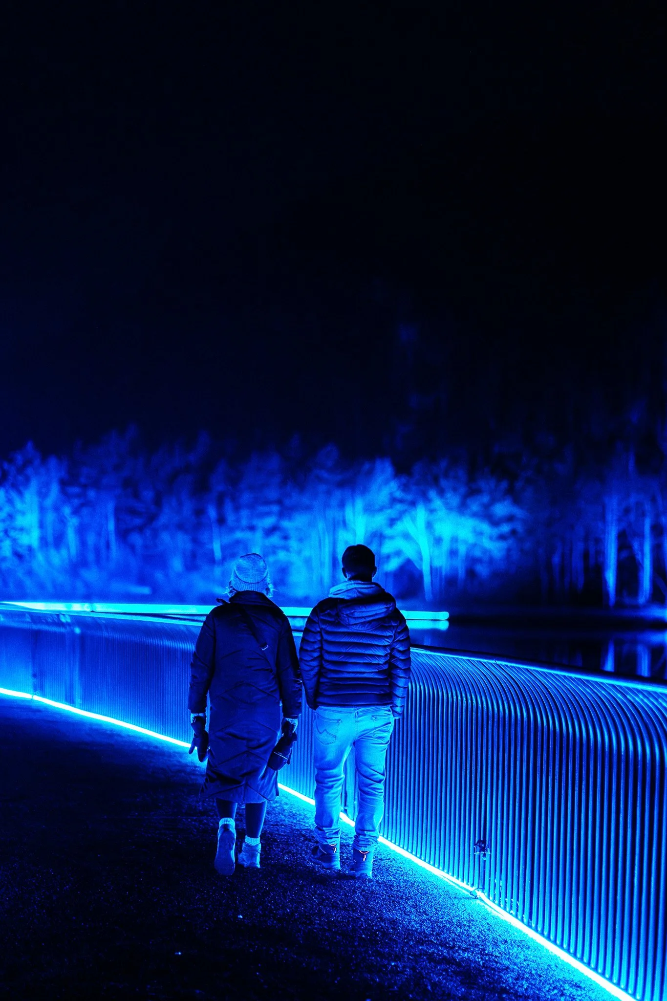 Twee mensen wandelen langs een verlichte, blauwe buisvormige structuur 's nachts, met een donkere hemel en bomen op de achtergrond.
