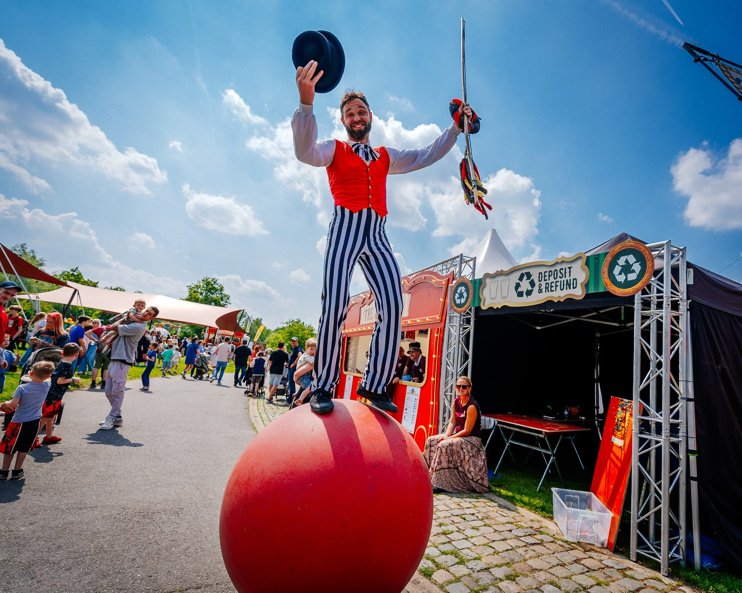 Man op grote rode bal als acrobatiek act tijdens een festival, met een publiek en standen omringd.