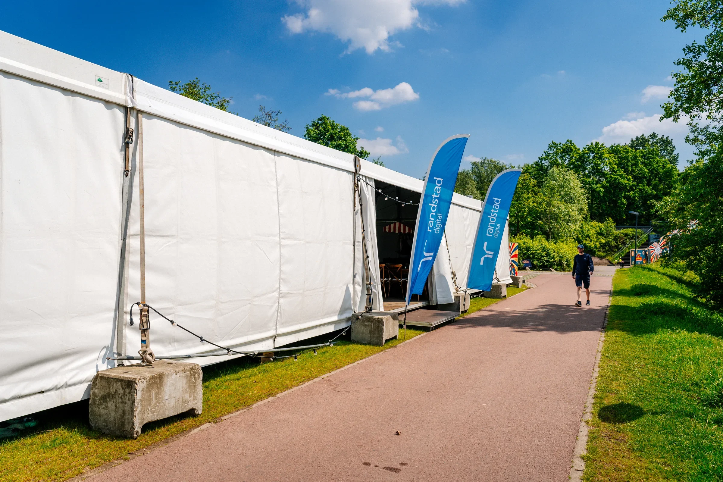 Een witte tent op een grasveld in een park met blauwe vlaggen die 'randsstad digital' zeggen, omgeven door groene bomen en een heldere blauwe hemel met enkele witte wolken. Mensen wandelen langs het pad naast de tent.