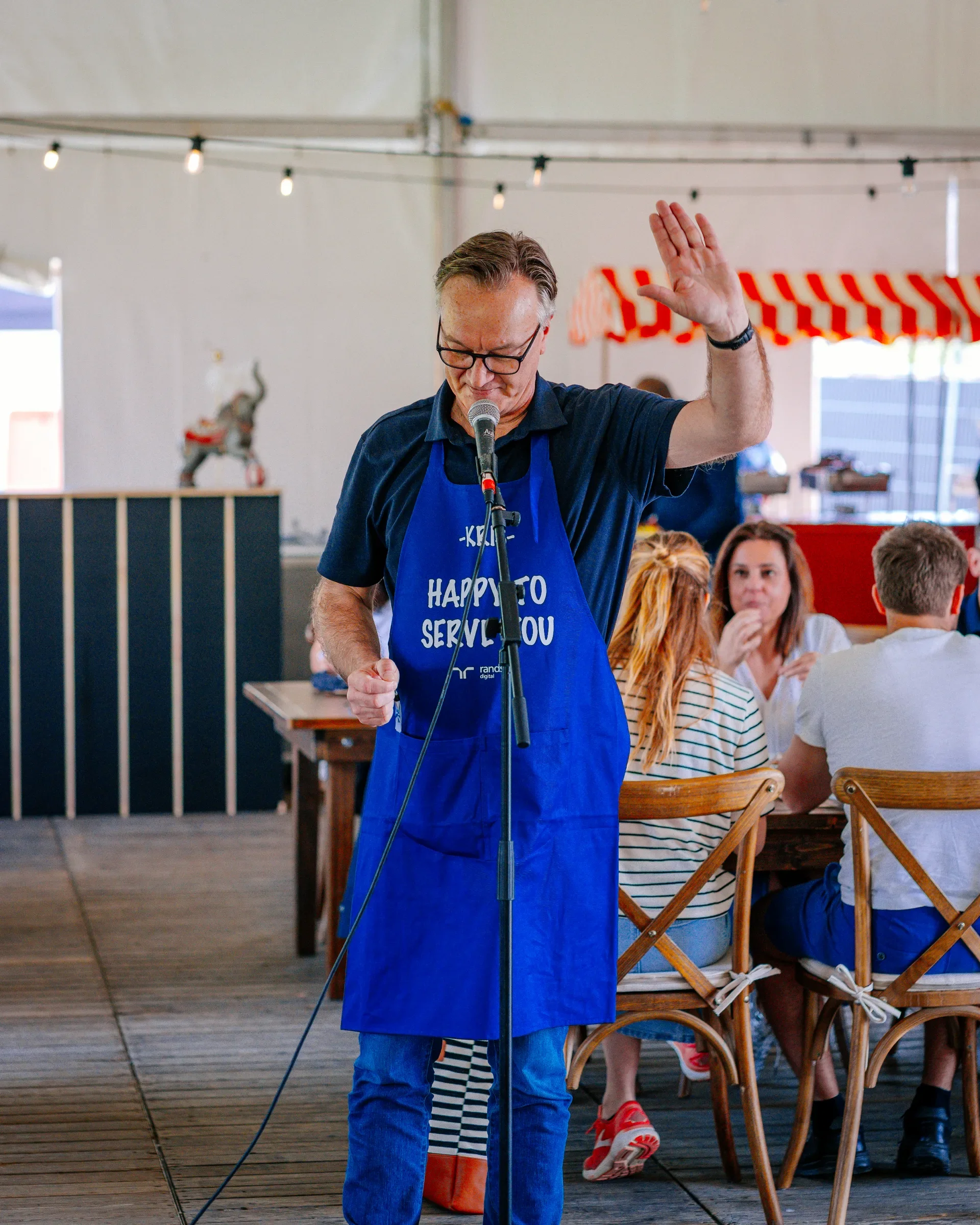 Man met bril en blauwe schort die bidden met hand omhoog in een restaurant of café.