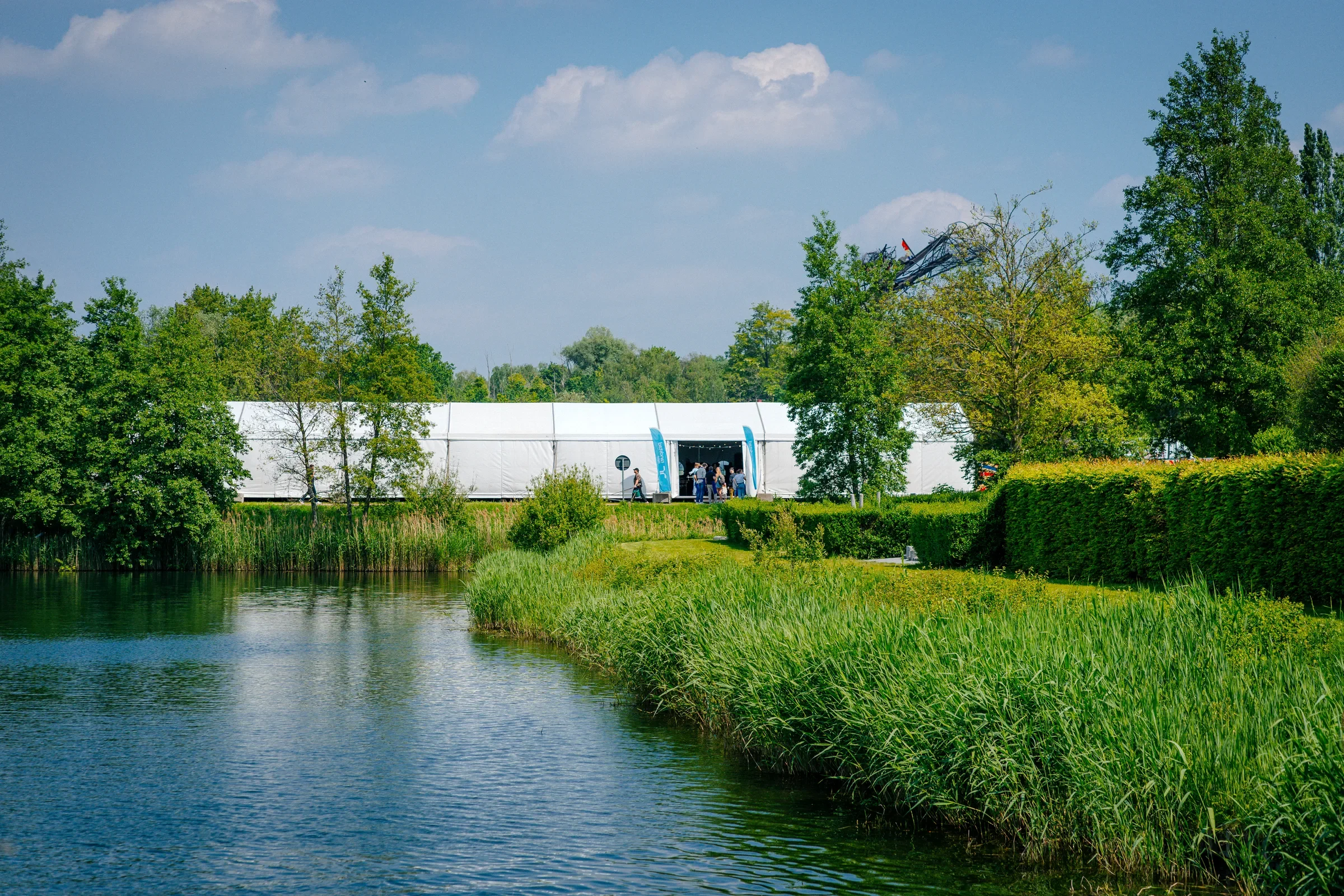 Een natuurlandschap met een rivier in de voorgrond, omringd door groene bomen en struiken, en een grote witte tent met mensen eronder op de achtergrond, met een blauwe lucht en enkele wolken.