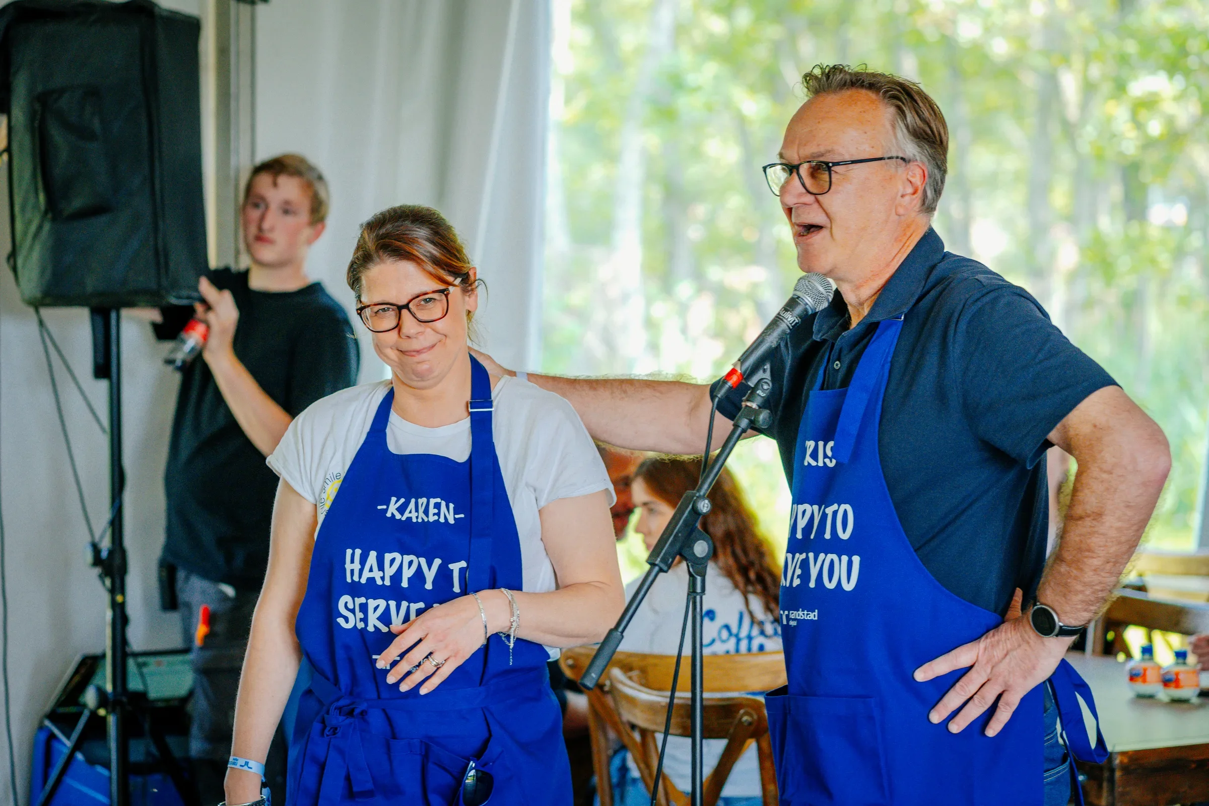 Mensen werken in een keuken tijdens een evenement, twee mensen dragen blauwe schorten met tekst, op de achtergrond een man met een microfoon en een andere persoon.