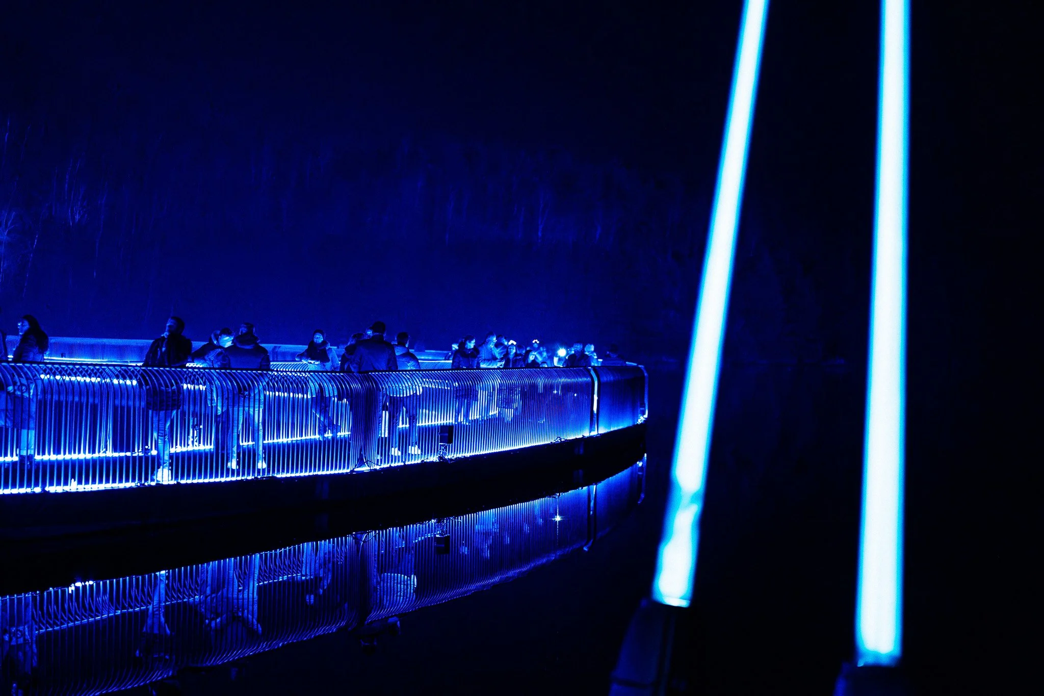 Een groep mensen staat op een brug of platform die verlicht is met blauwe neonlichten, 's nachts.