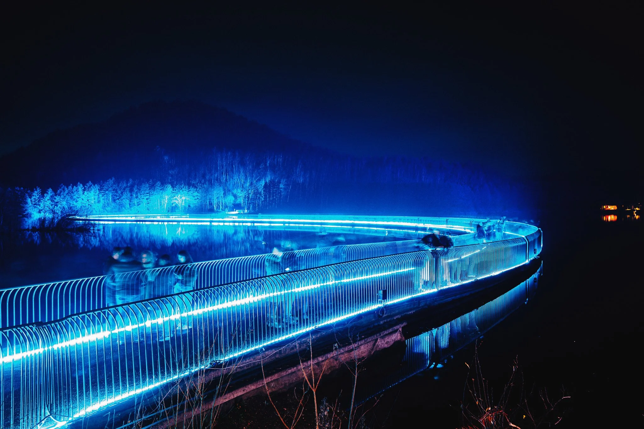Verlichte wandelroute met blauwe LED-verlichting langs een meer, omgeven door bergachtige landerijen in de nacht.