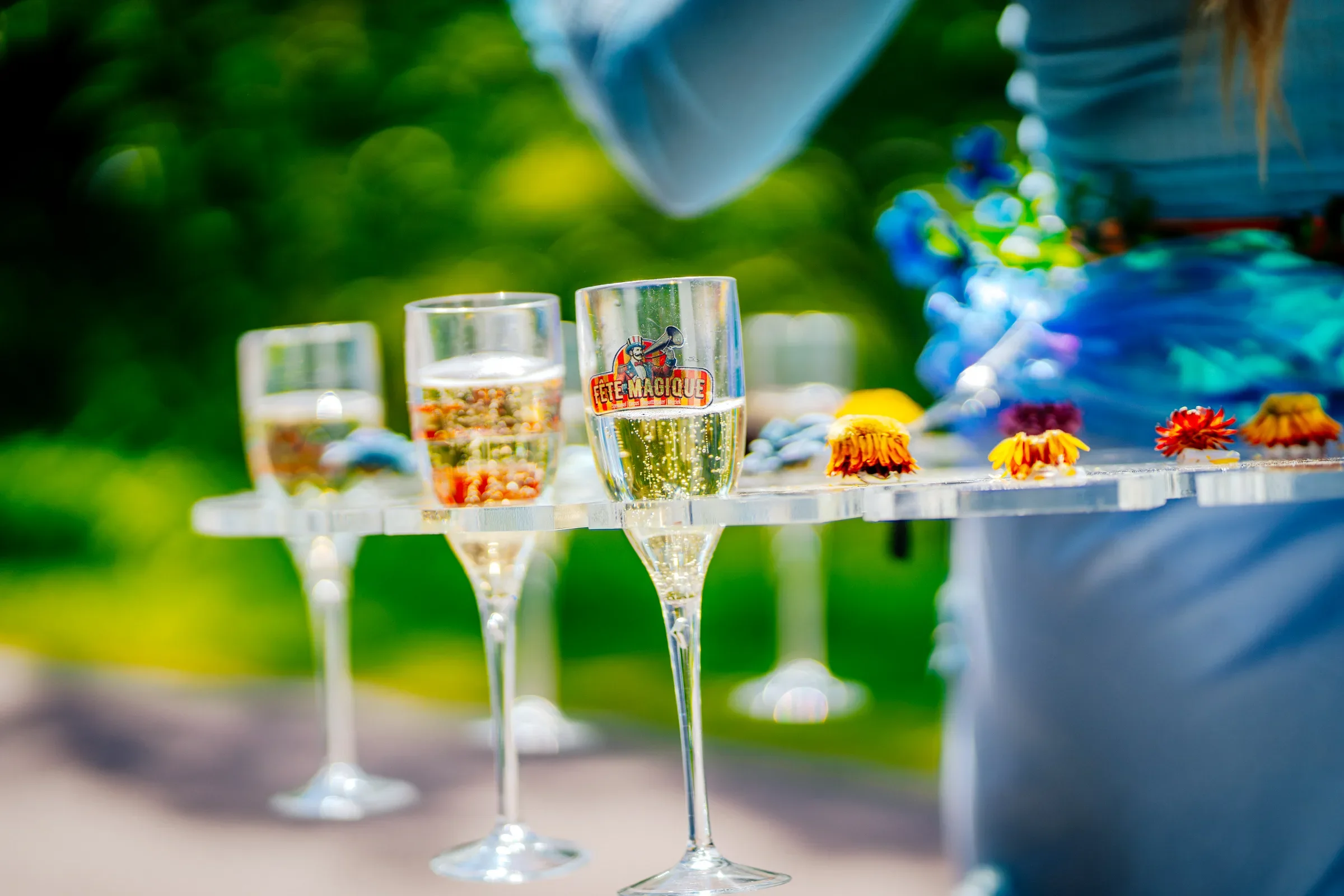 Twee glazen met champagne, een met een sticker die zegt 'Fête Magique', en kleurrijke bloemen op een tafel, buiten met groene achtergrond.