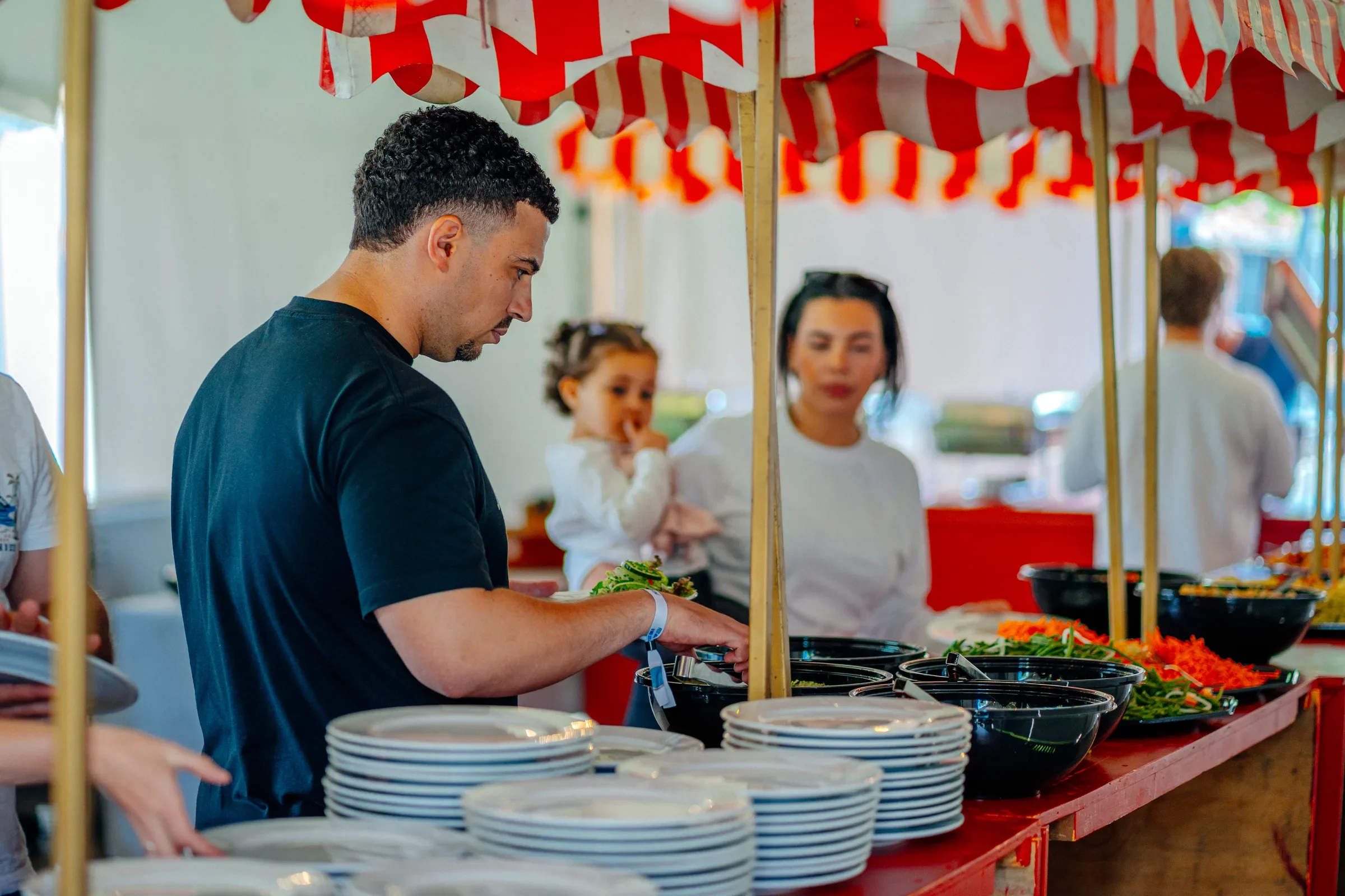 Vrouw en klein meisje bij een buffet met salades en groenten, onder een rode en witte parasol in een openlucht marktsituatie.