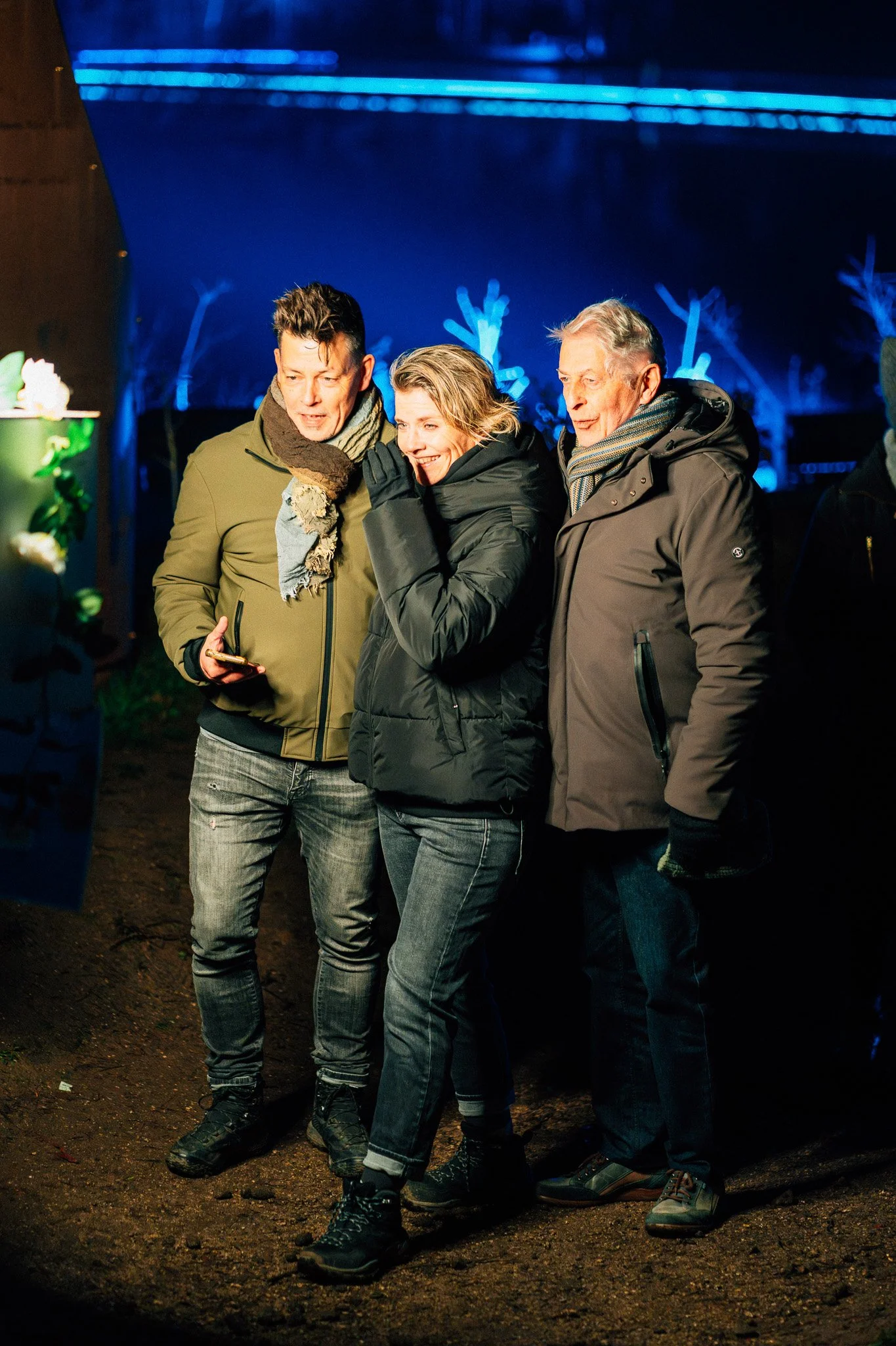 Drie mensen die buiten in de avond op het strand staan, gekleed in warme jas en sjaal, samen naar iets kijken en plezier hebben.