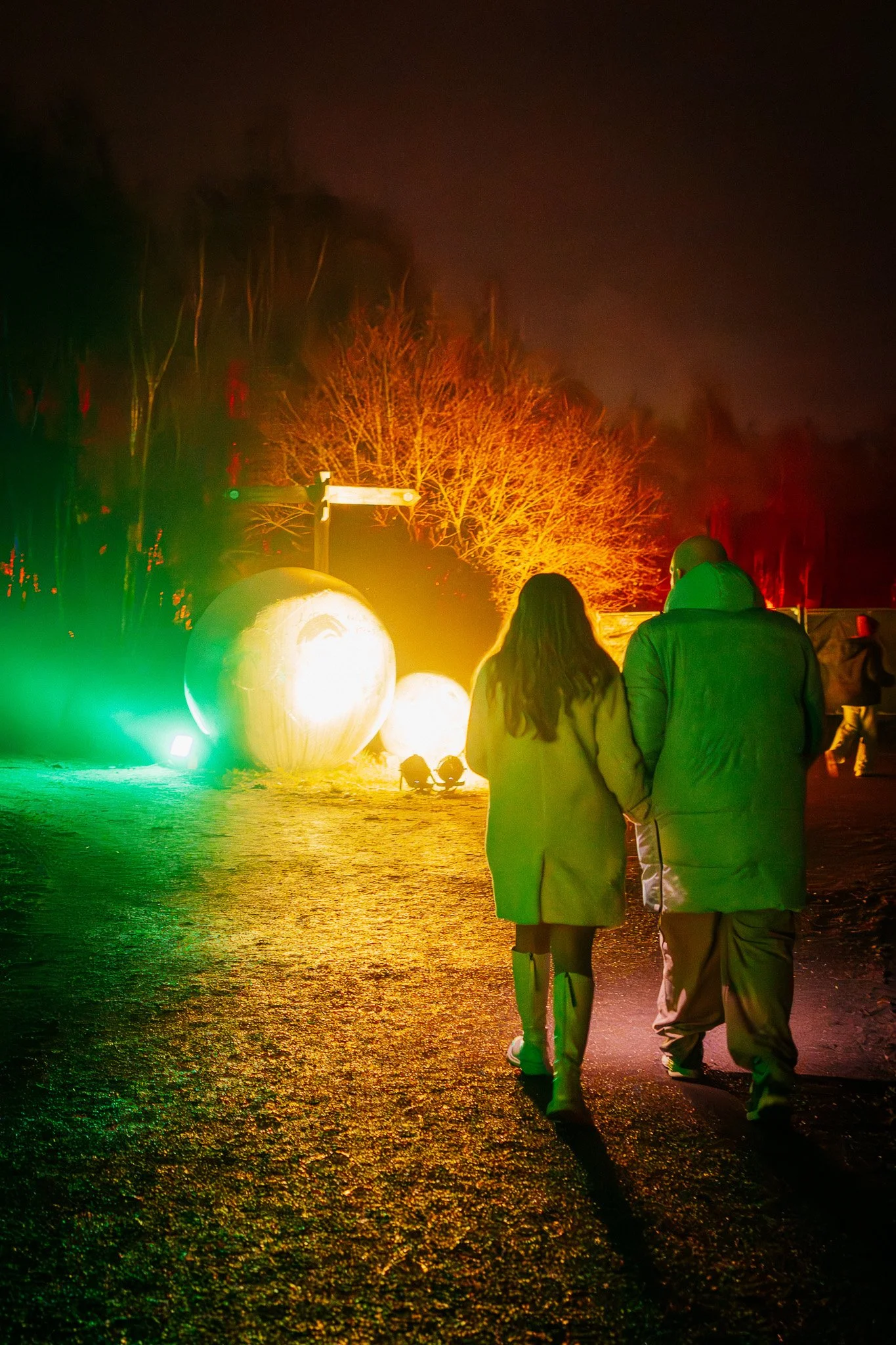 Een paar wandelaars lopen hand in hand op een verlichte straat 's nachts, met ballonnen en kleurrijke feestverlichting op de achtergrond.
